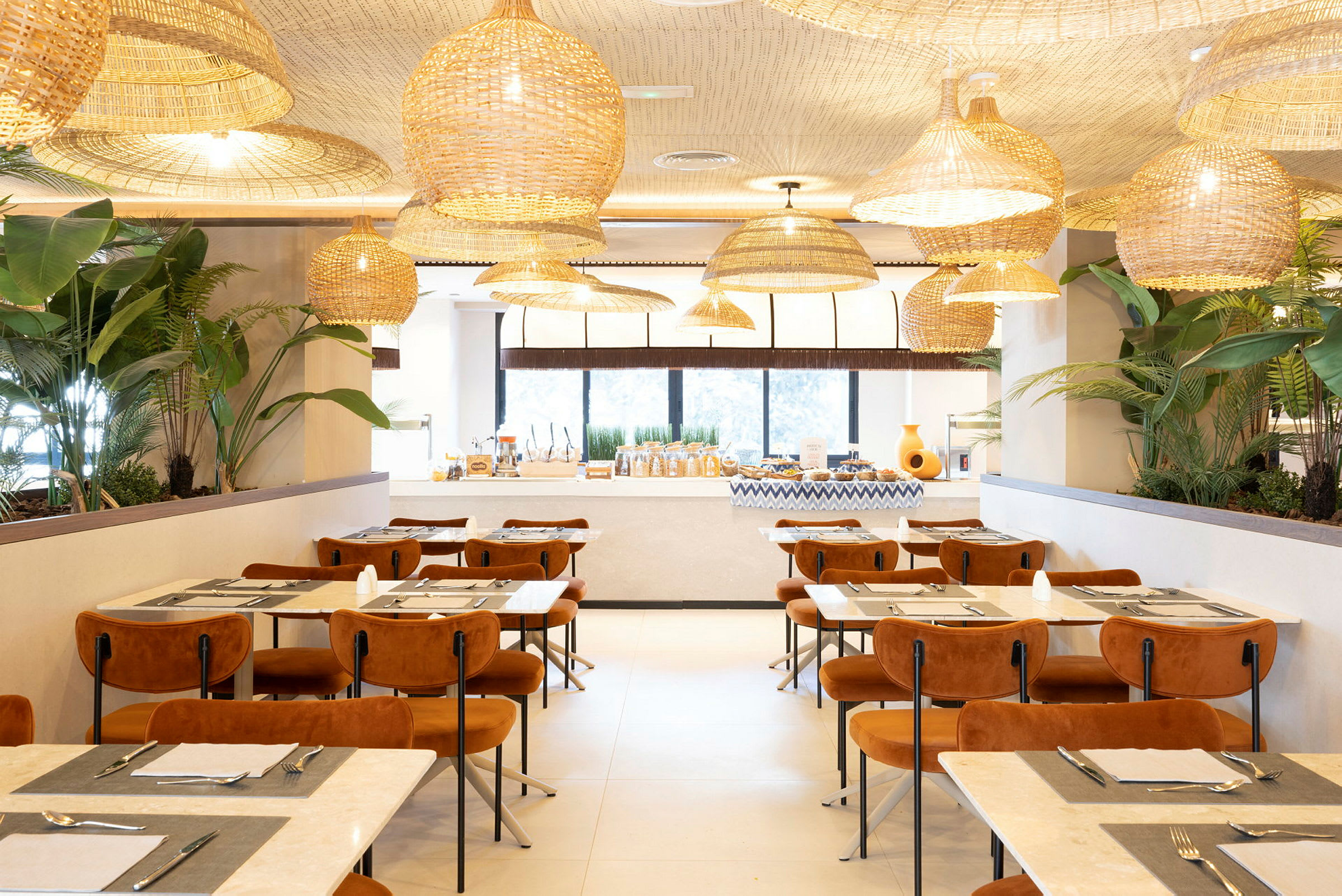 En elegant restaurant med moderne møbler, store vinduer, planter og hengende kurvlamper i taket. (AI-generert tekst)