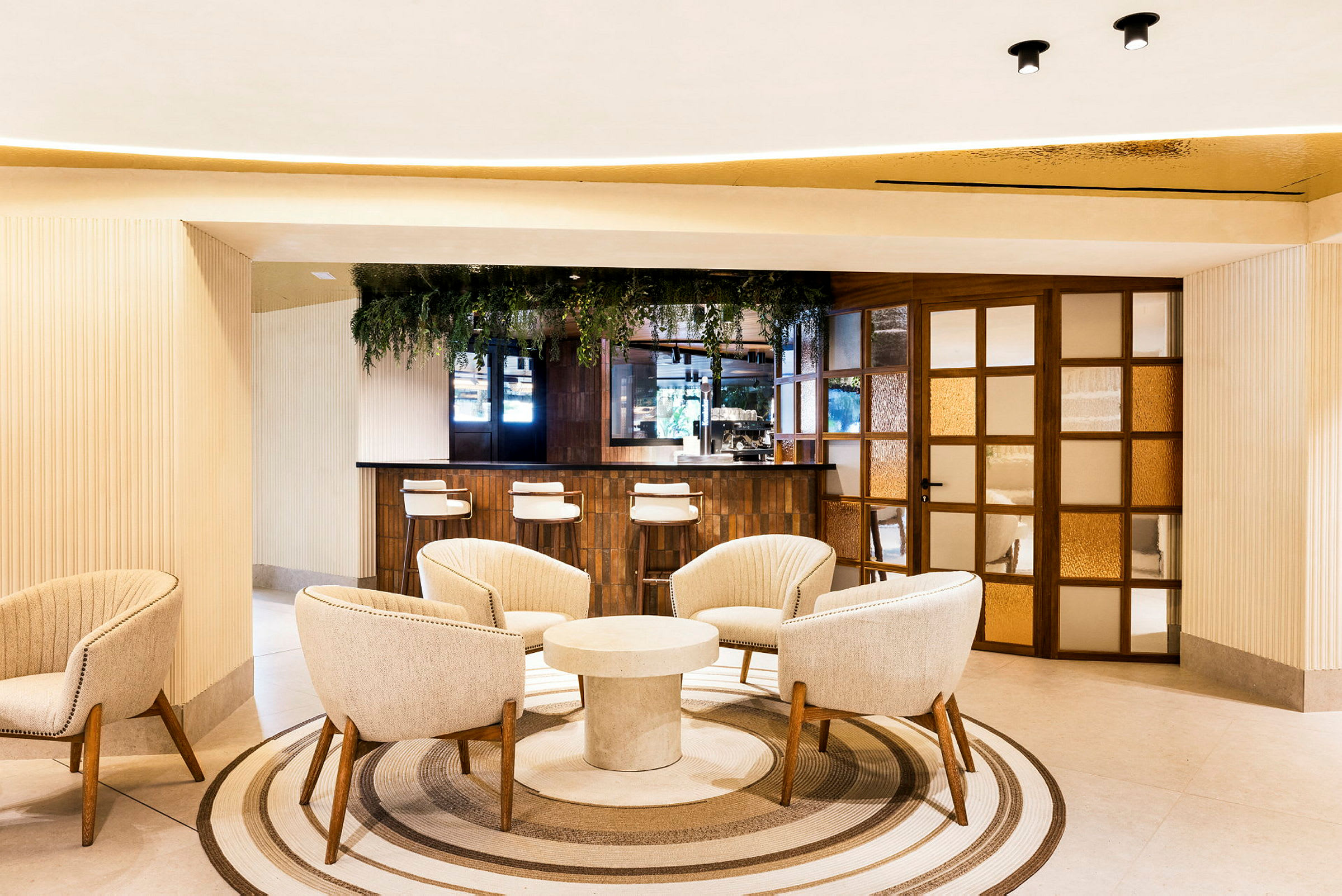 Et moderne hotellområde med en sittegruppe med myke lenestoler rundt et rundt bord og en bardisk i bakgrunnen. Innredningen er lys og elegant med plantepynt. (AI-generert tekst)