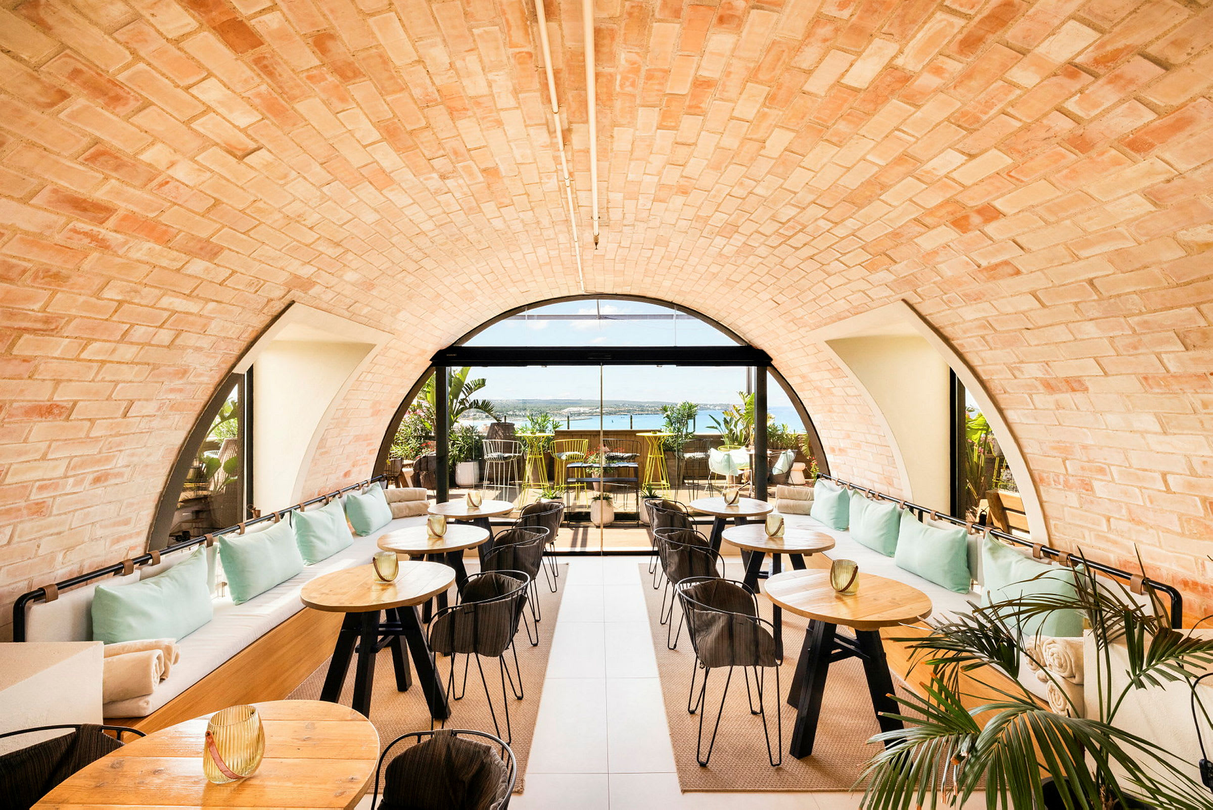 En stilfull kafé med teglmurtak og store vinduer som vender ut mot en terrasse med utsikt over havet. Bordene er tre med svarte ben, og det finnes dekorative planter. (AI-generert tekst)