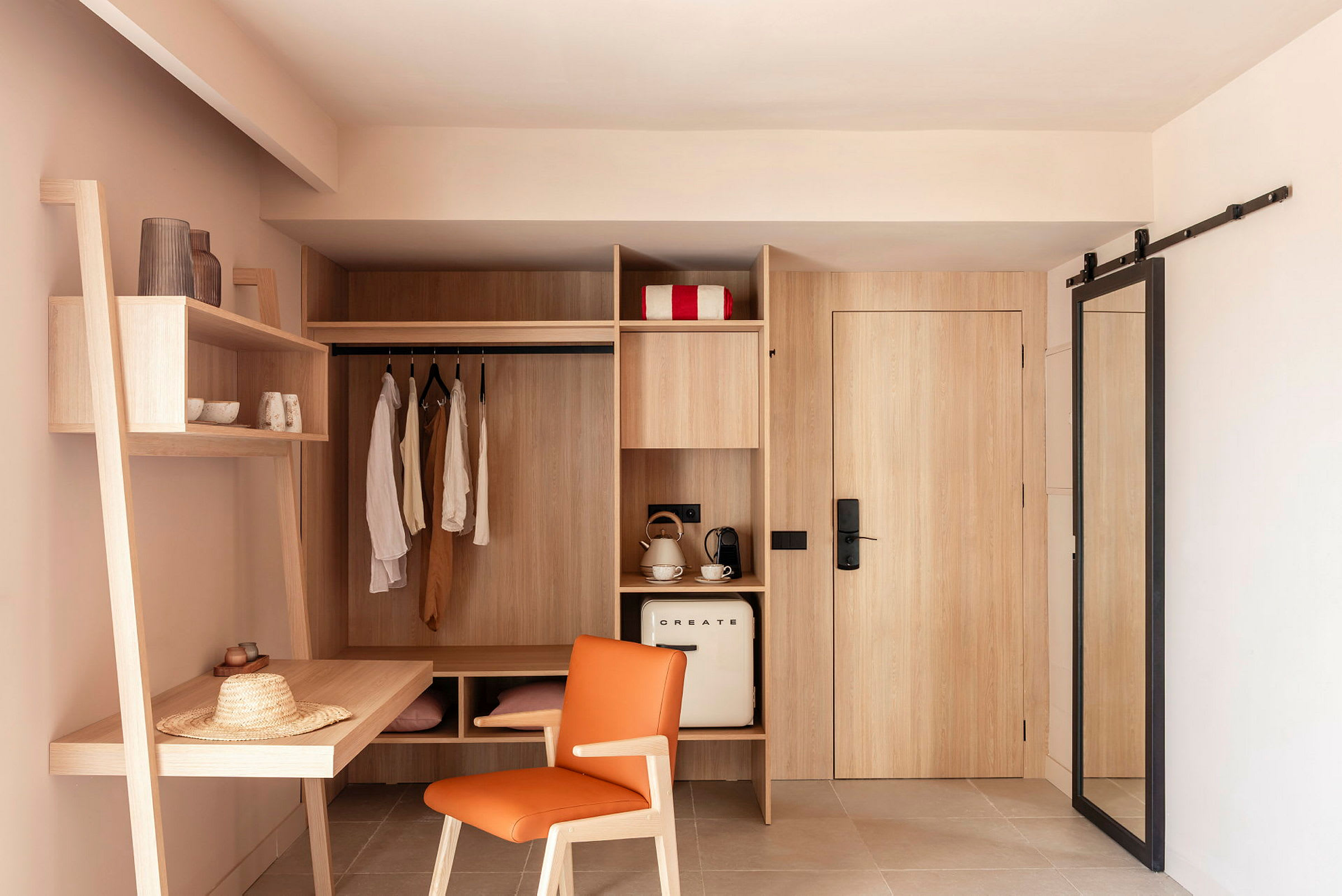 Bildet viser et hotellrom med innebygget oppbevaringsplass, inkludert kroker, hyller og et skrivebord. Et kjøleskap, en kaffemaskin og en vannkoker er også synlige. (AI-generert tekst)