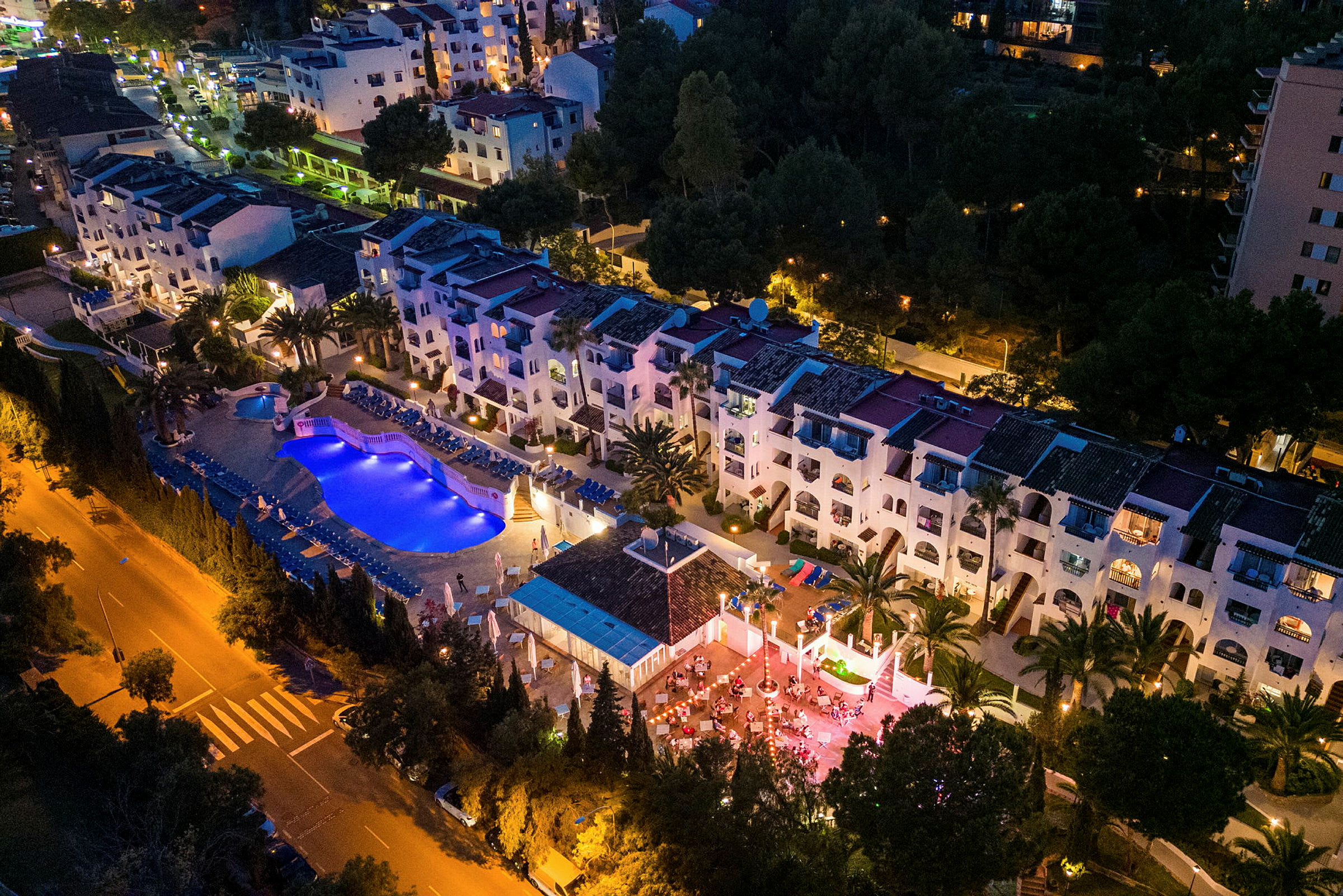 Flyfoto av et opplyst hotellanlegg med basseng og terrasse om kvelden. (AI-generert tekst)