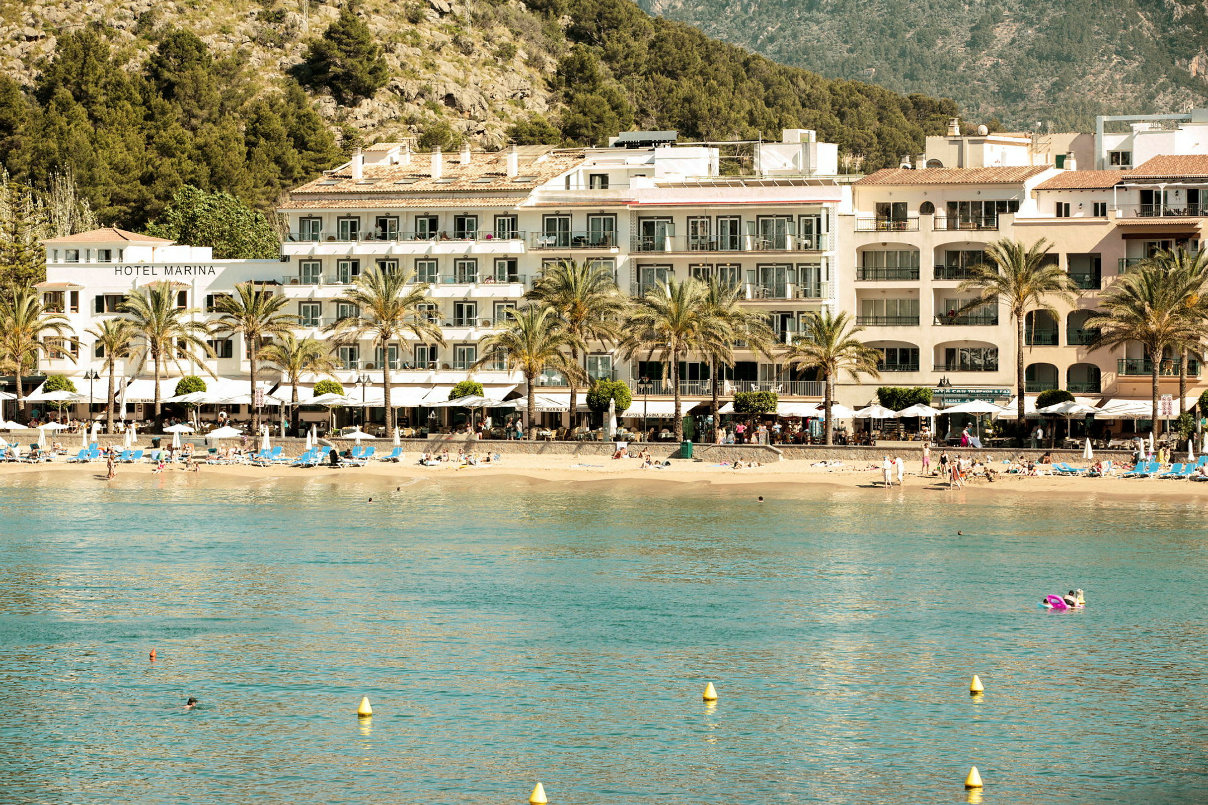 Hotell og strand ved kysten med fjell og trær i bakgrunnen. (AI-generert tekst)