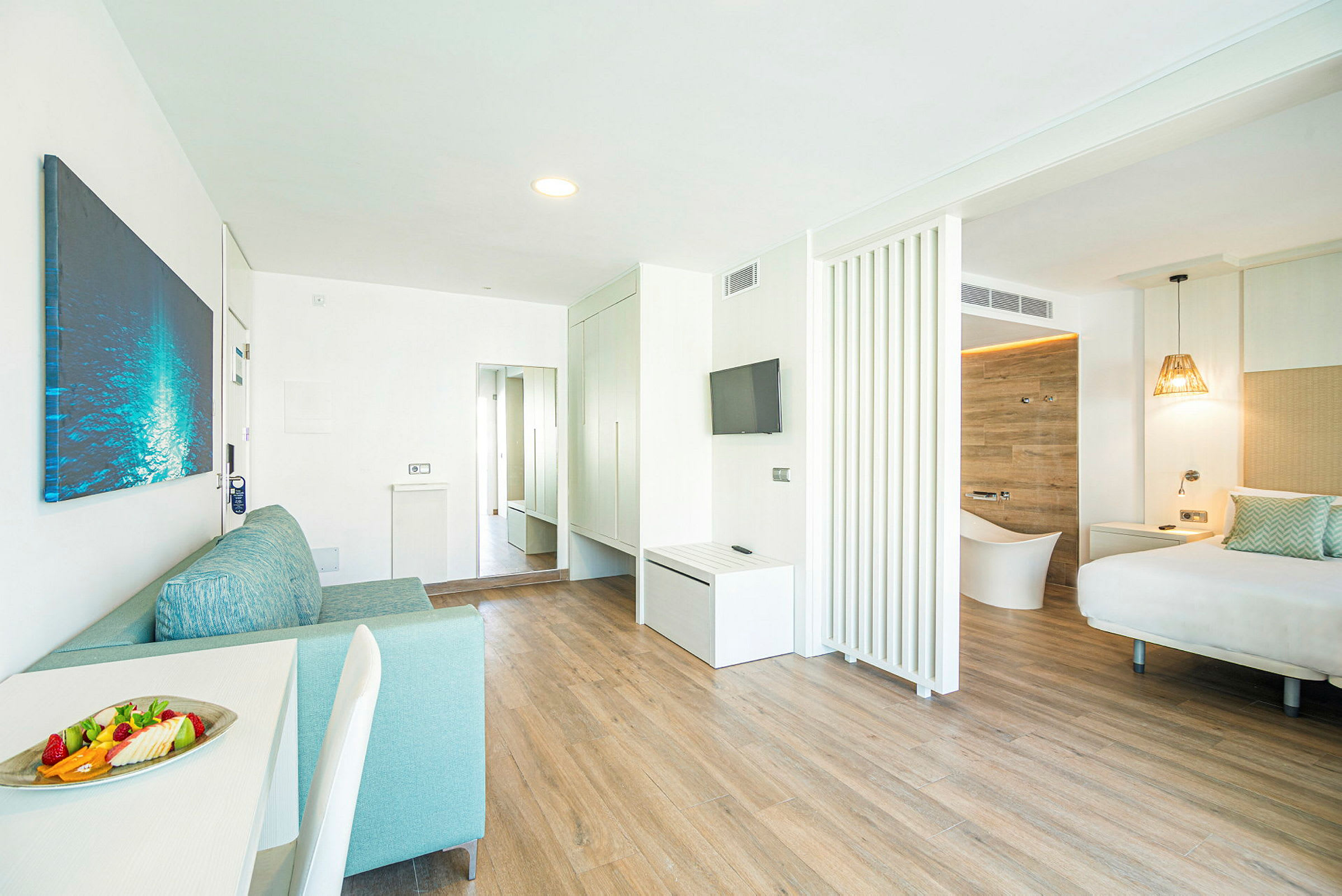 Et moderne hotellrom med lys innredning, tregulv og separate sove- og oppholdsromsområder. En tallerken med frukt er plassert på bordet. (AI-generert tekst)