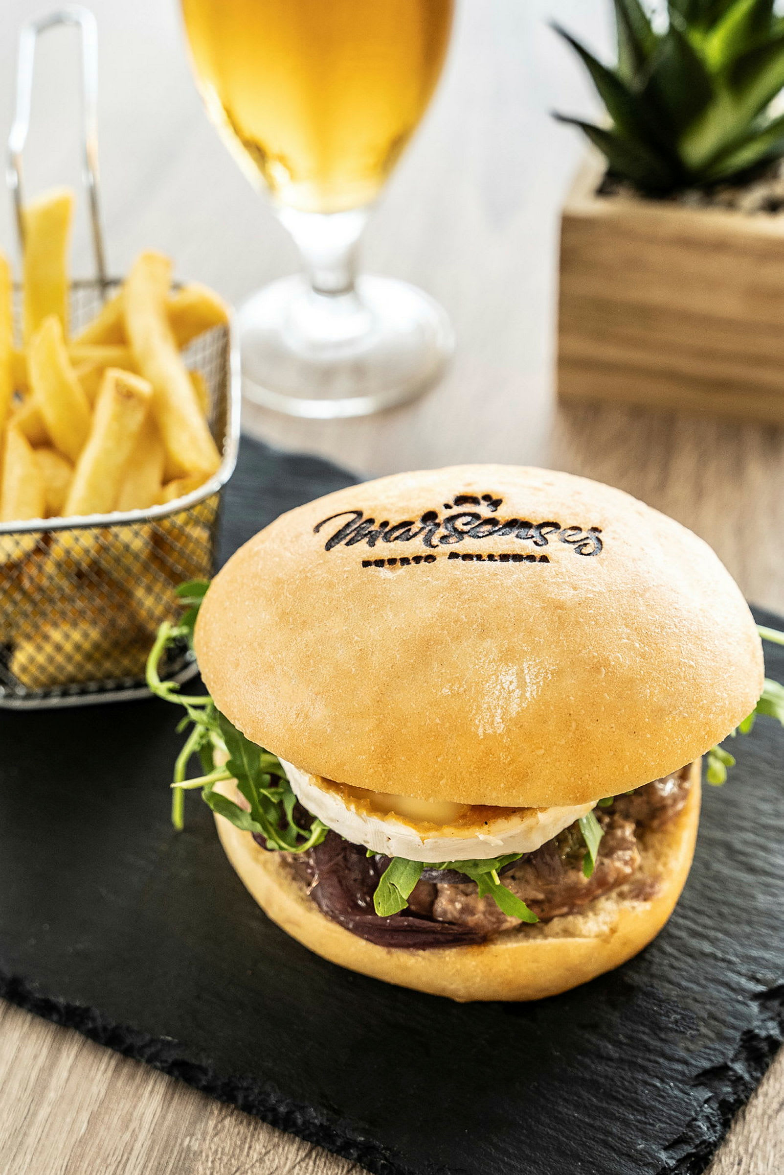 Nærbilde av en hamburger servert med pommes frites og et glass øl i bakgrunnen. (AI-generert tekst)