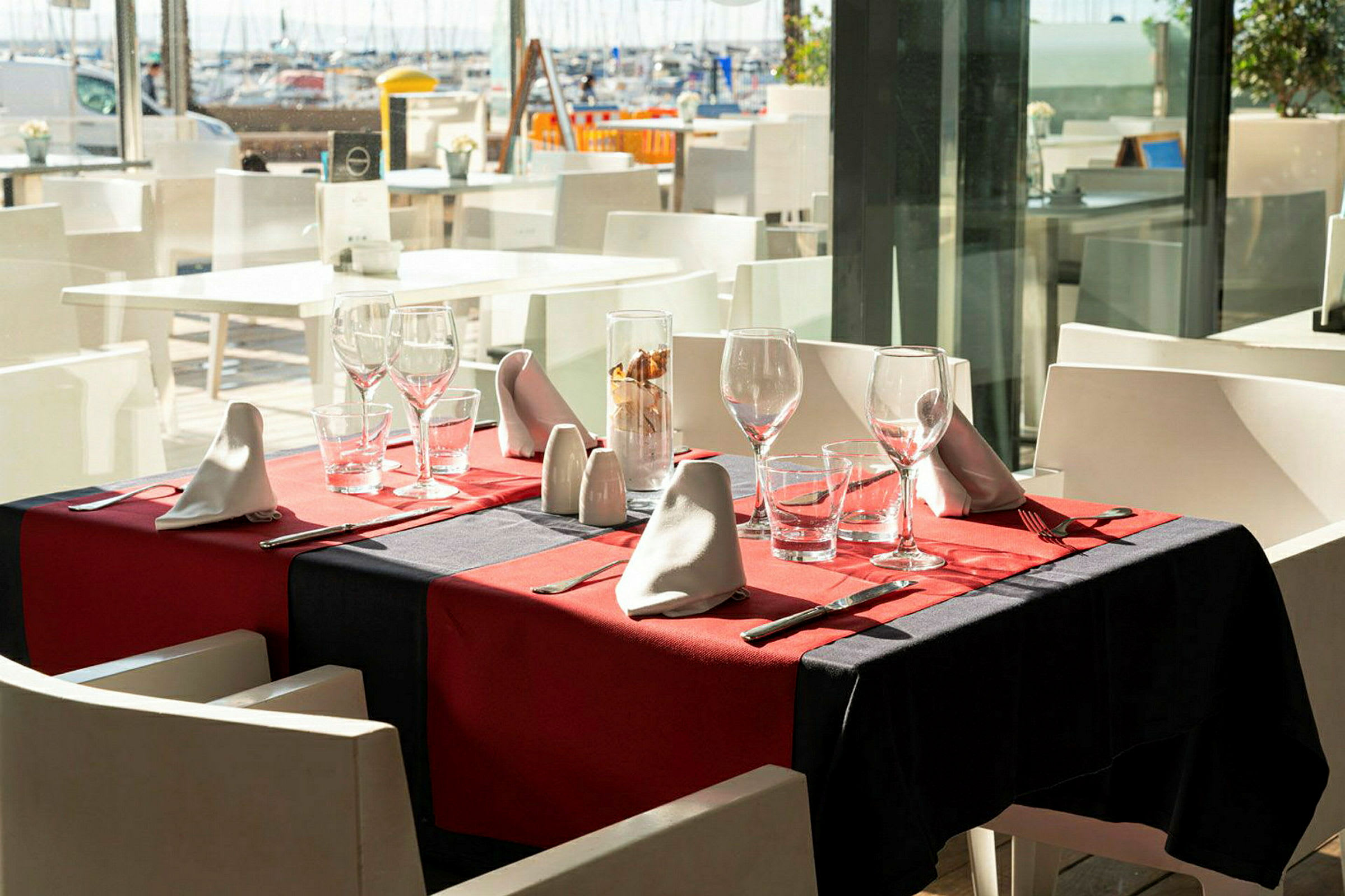 Et dekket bord med rød duk, glass og bestikk på en restaurant med utsikt over havnen. (AI-generert tekst)