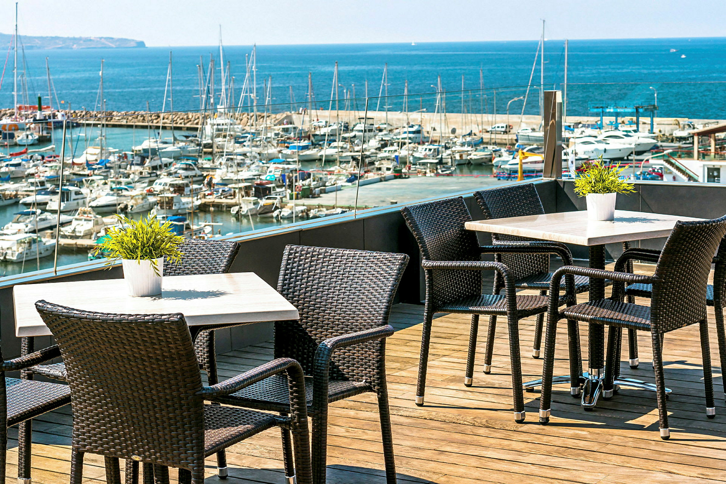Uteservering med bord og stoler på en terrasse med utsikt over en småbåthavn og havet. (AI-generert tekst)