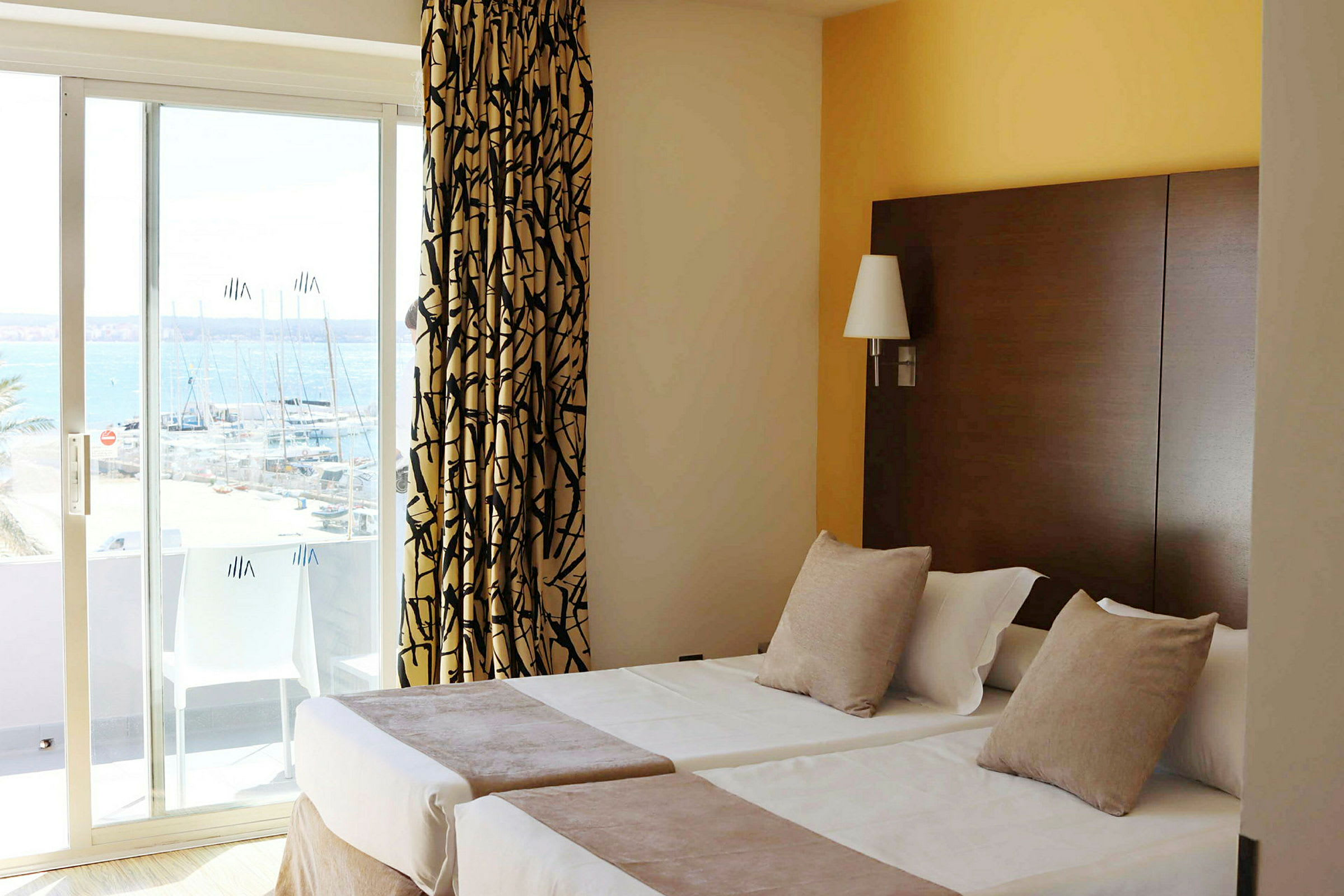 Hotellrom med to enkeltsenger, dekorert med puter og sengeteppe. Et vindu med skyvedør viser utsikt over en marina. (AI-generert tekst)