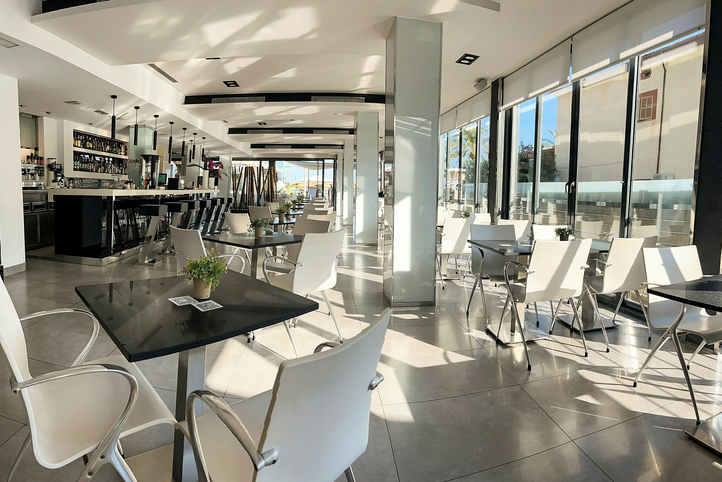 En moderne restaurant med hvite stoler og bord, samt en lang bar til venstre. (AI-generert tekst)
