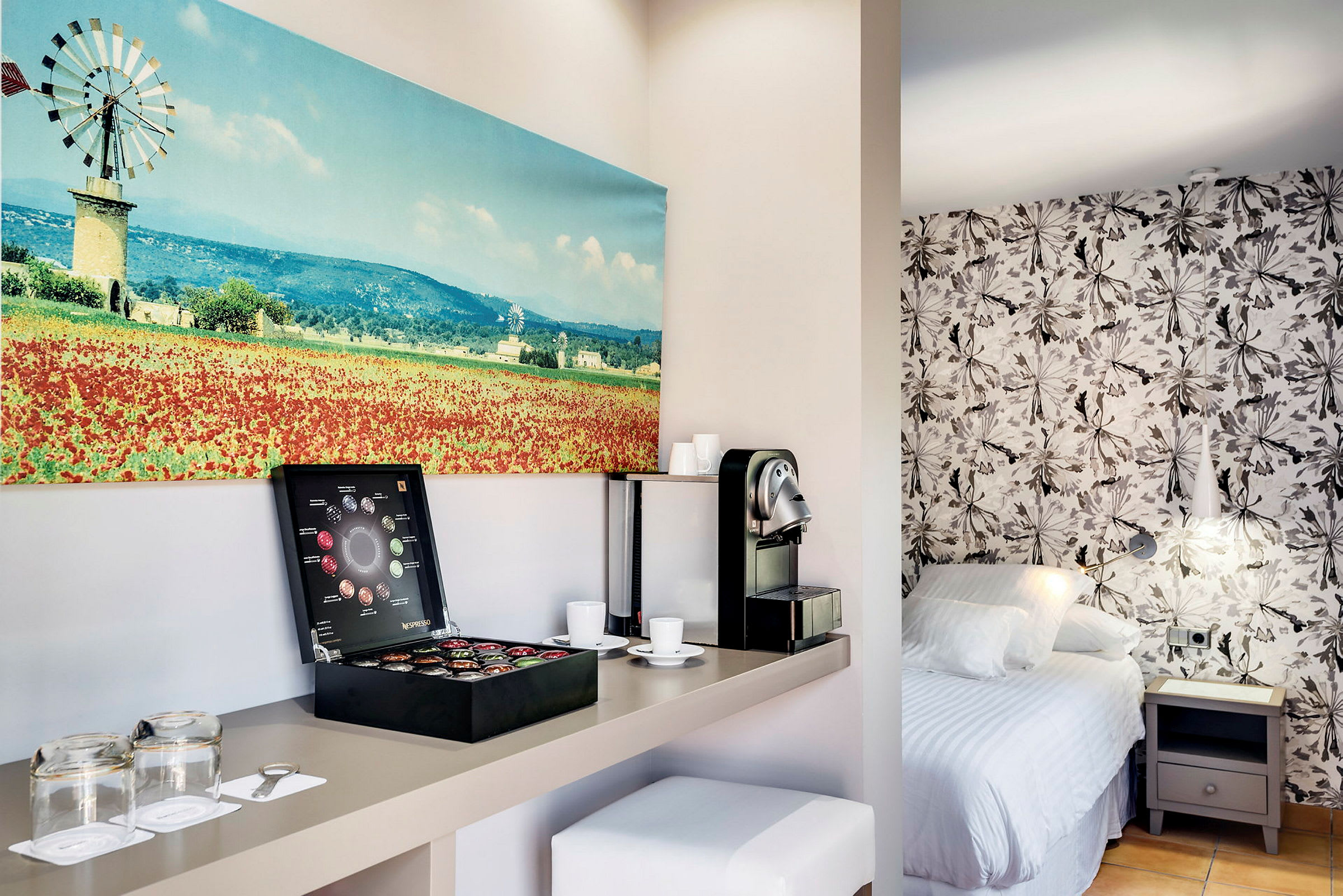 Et hotellrom med en kaffemaskin og kaffekapsler på en hylle, en seng med en blomstrete tapet og et maleri av et landskap. (AI-generert tekst)