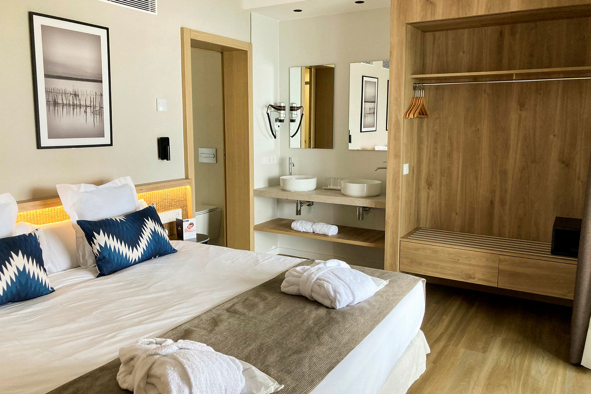Et hotellrom med en dobbeltseng, to blå puter og hvite badekåper på sengen. I bakgrunnen er det et bad med to servanter og speil samt en garderobe med trepanel. (AI-generert tekst)