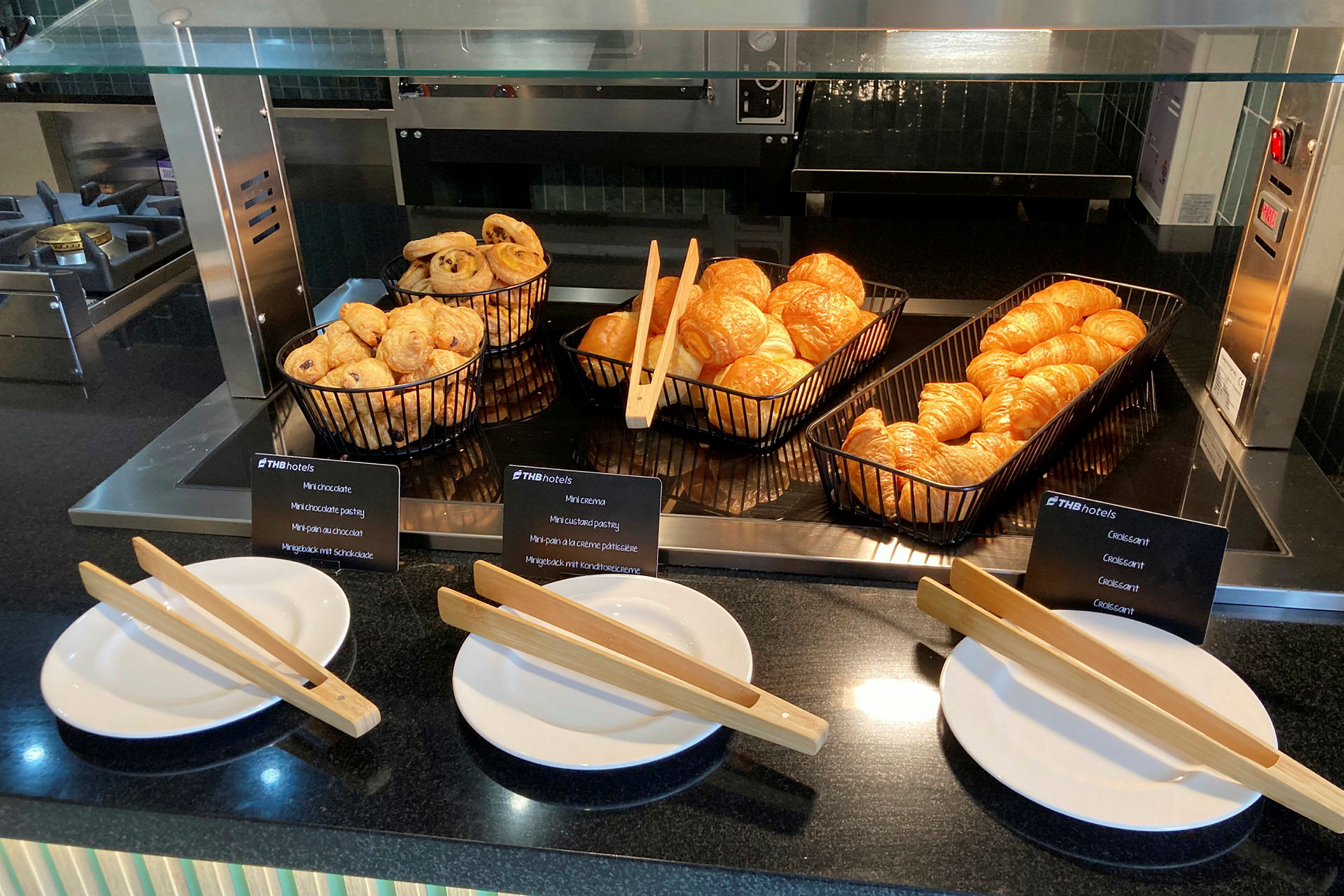 Frokostbuffé med ferske croissanter og bakverk i svarte kurver. Foran står hvite tallerkener med trepincetter. (AI-generert tekst)