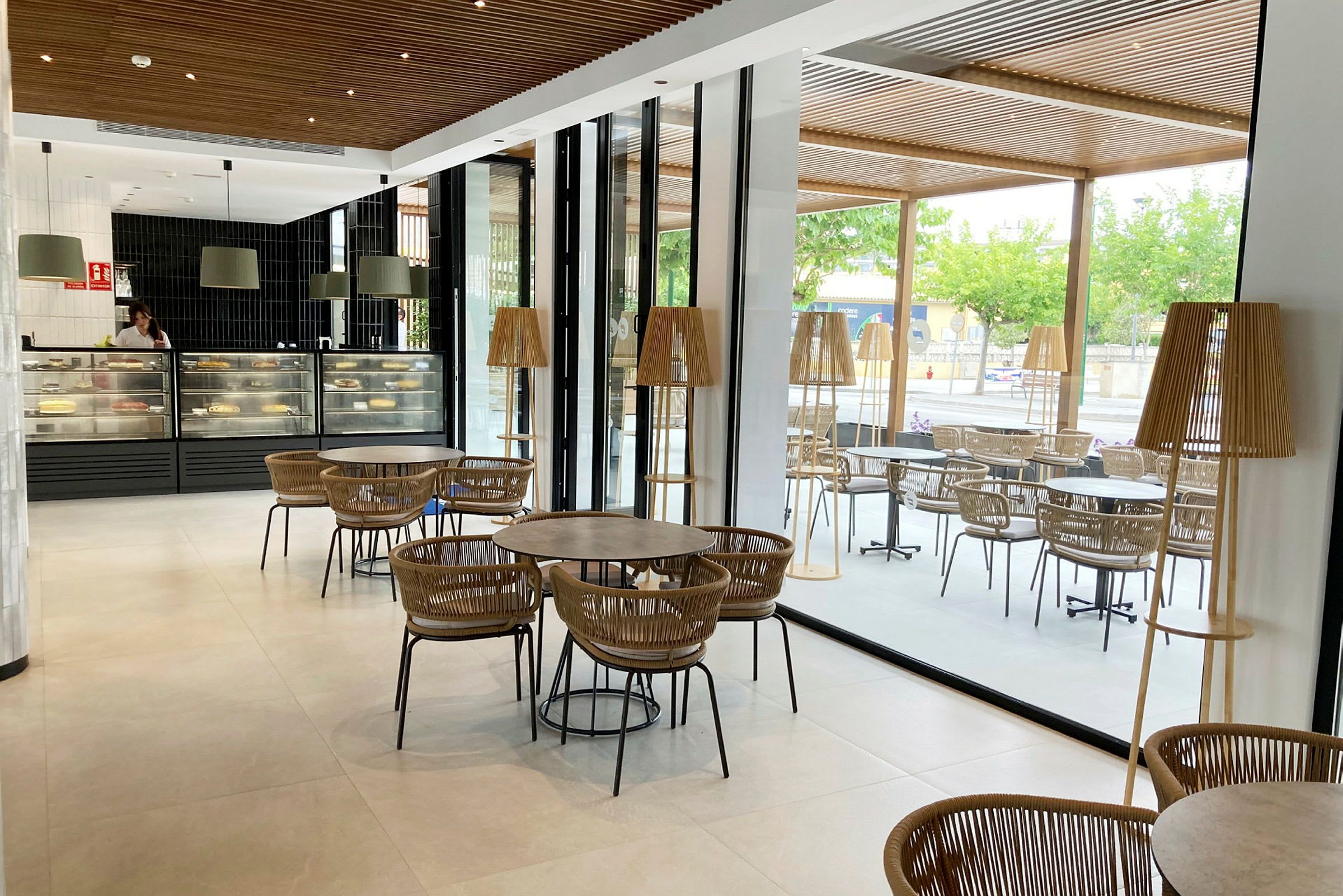En kafé med moderne møbler, store vinduer og glassvegger er vist på bildet. Det er flere bord og stoler samt en disk med bakverk. (AI-generert tekst)