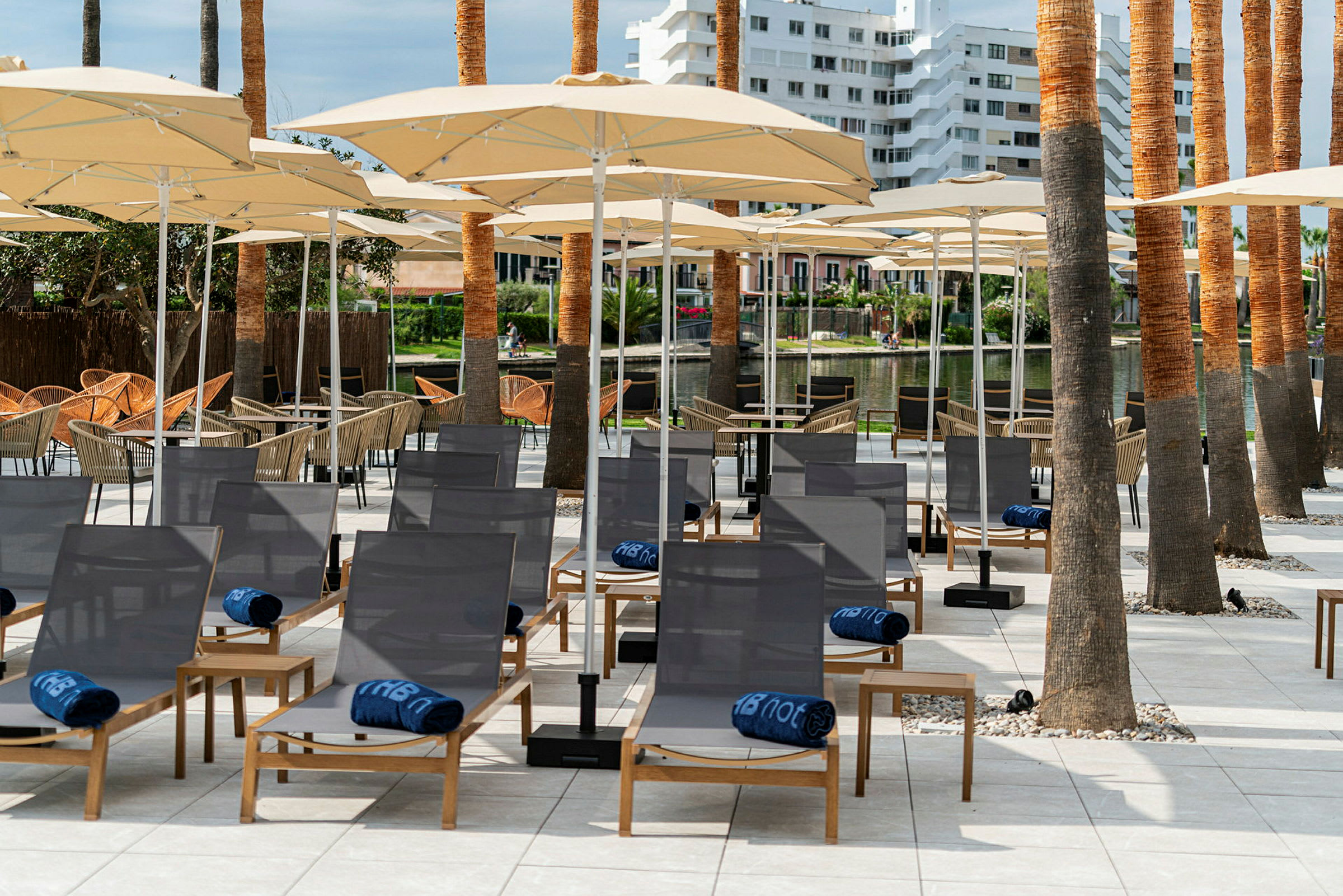 Solstoler med blå håndklær og parasoller på en terrasse omgitt av palmer. (AI-generert tekst)