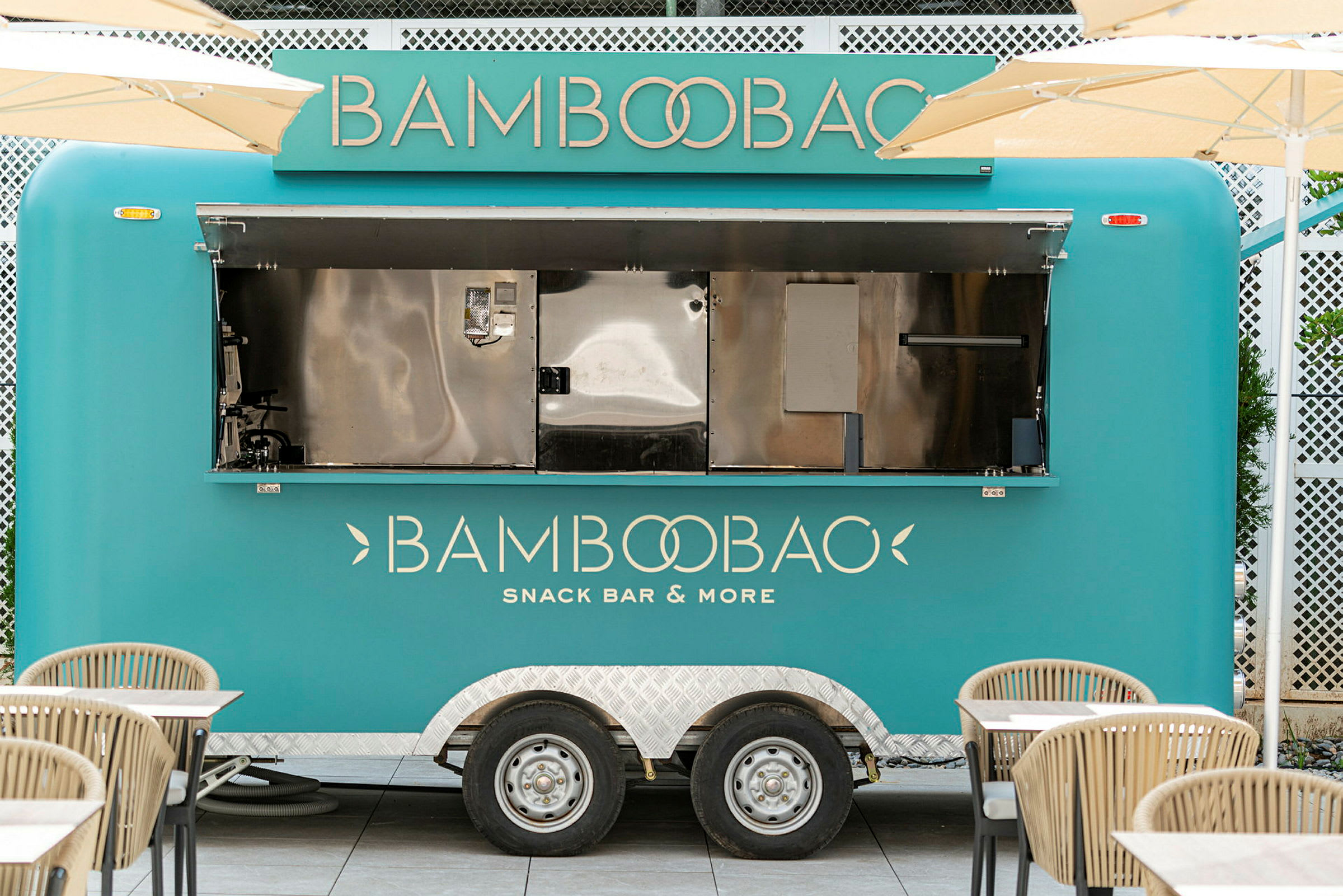 En turkis matvogn med teksten 'Bamboobao Snack Bar & More' står parkert omgitt av bord og stoler. (AI-generert tekst)