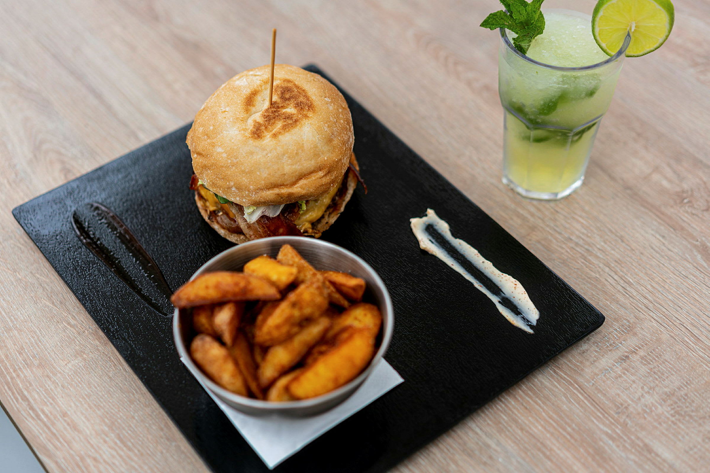 En matrett bestående av en hamburger, en bolle med båtskjærte poteter og en forfriskende drikke med lime og mynte. (AI-generert tekst)