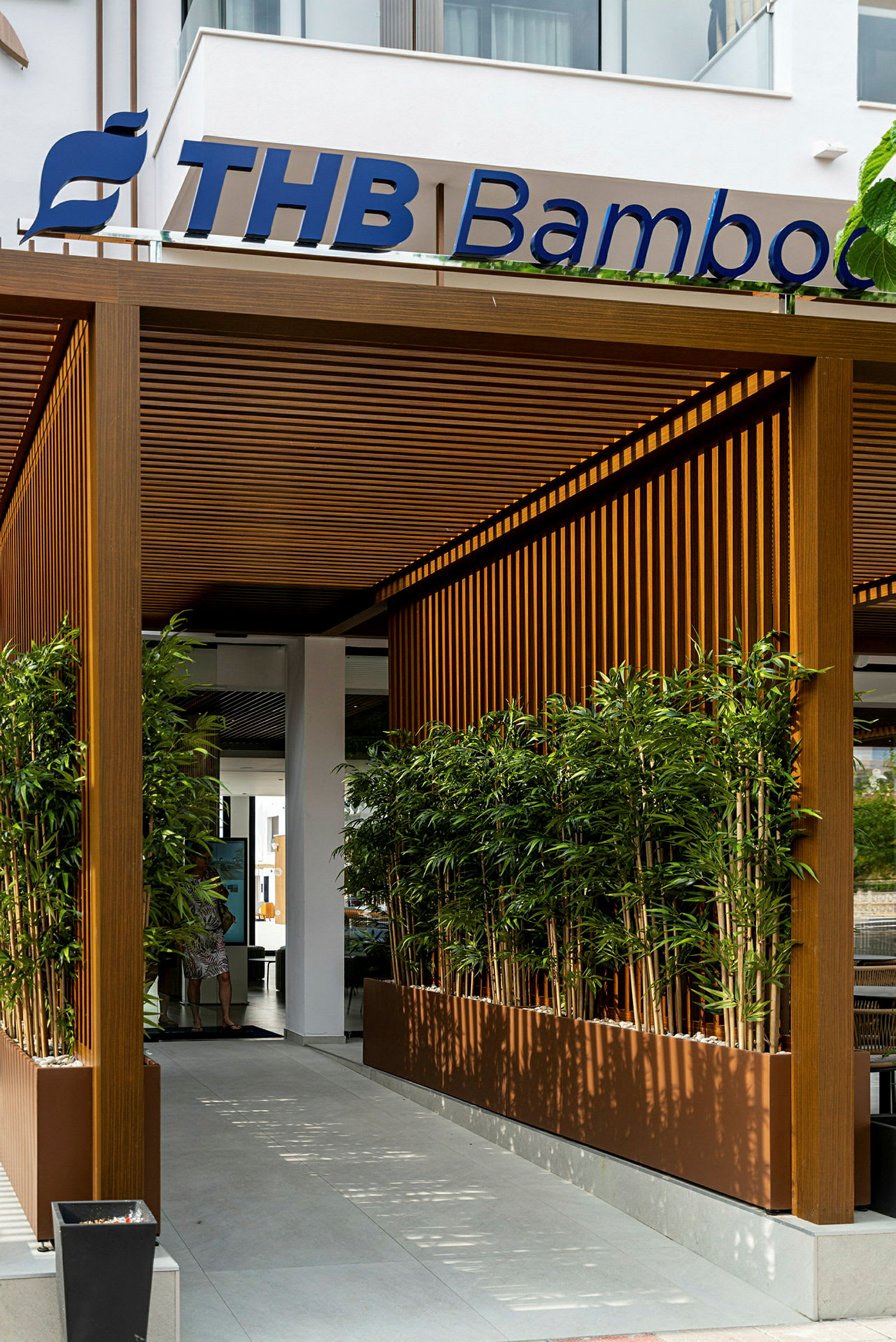 Inngangen til en bygning med en pergola i tre, omgitt av bambusplantinger. (AI-generert tekst)