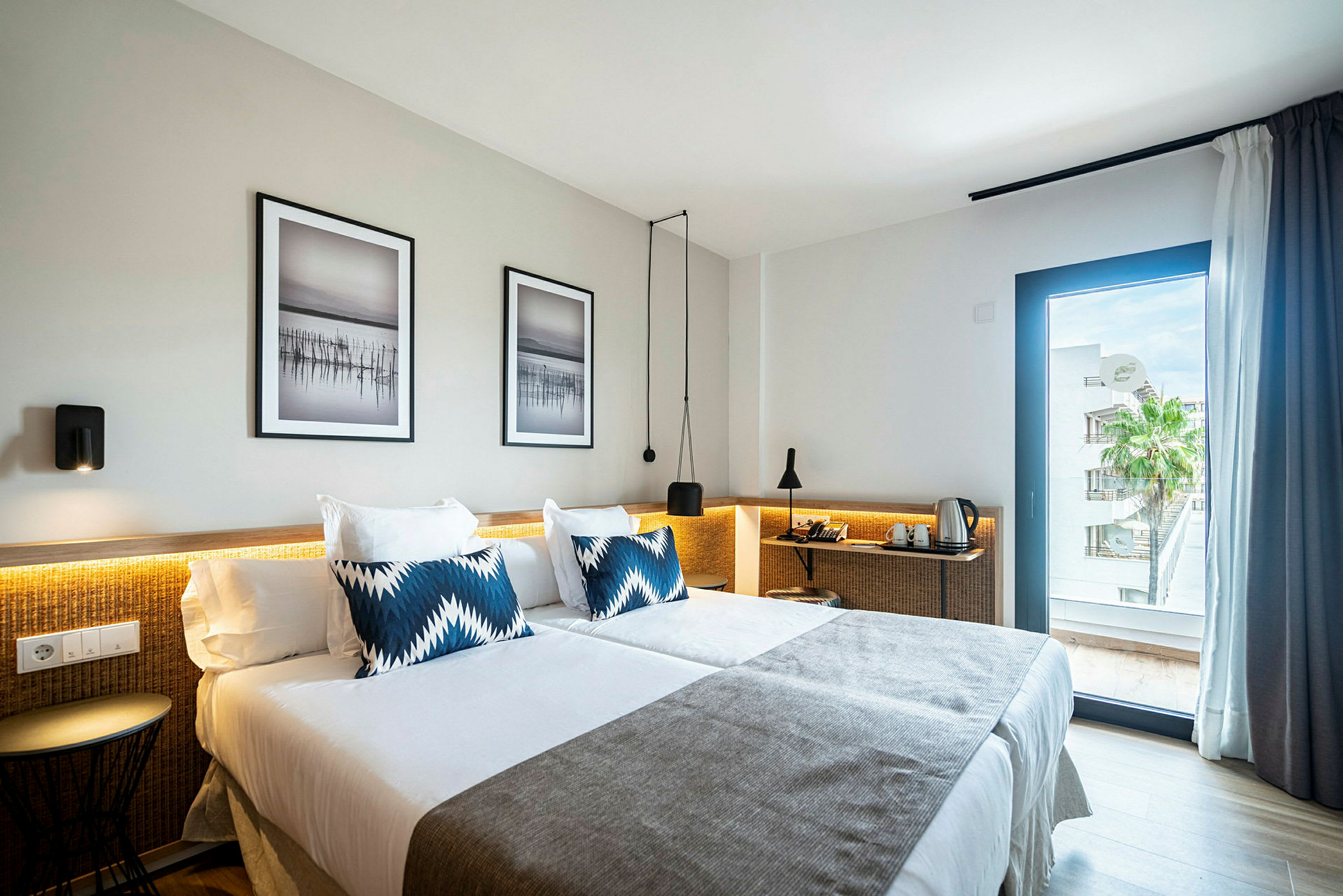 Et hotellrom med to senger dekket av hvitt sengetøy og grå sengetepper. Det er blå dekorative puter på sengene. Veggene har to innrammede kunstbilder, og et vindu viser utsikt over en palme og bygninger. (AI-generert tekst)