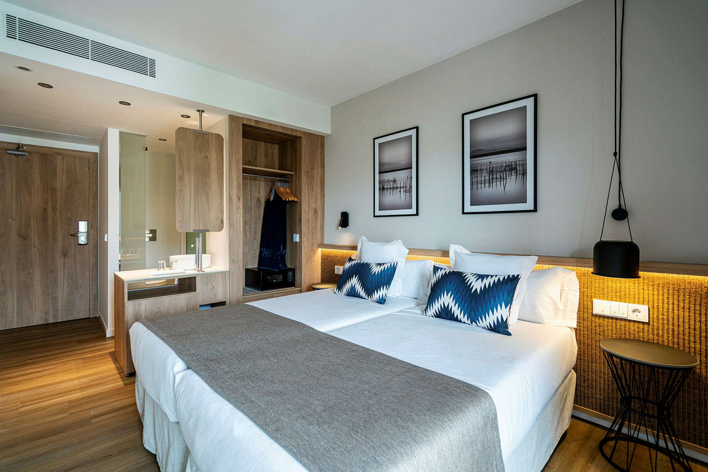 Et hotellrom med en stor dobbeltseng, beige sengeteppe og blåhvite puter. Veggen har to innrammede bilder over sengen, og til venstre er det en trebjelke med garderobe og skrivebord. (AI-generert tekst)