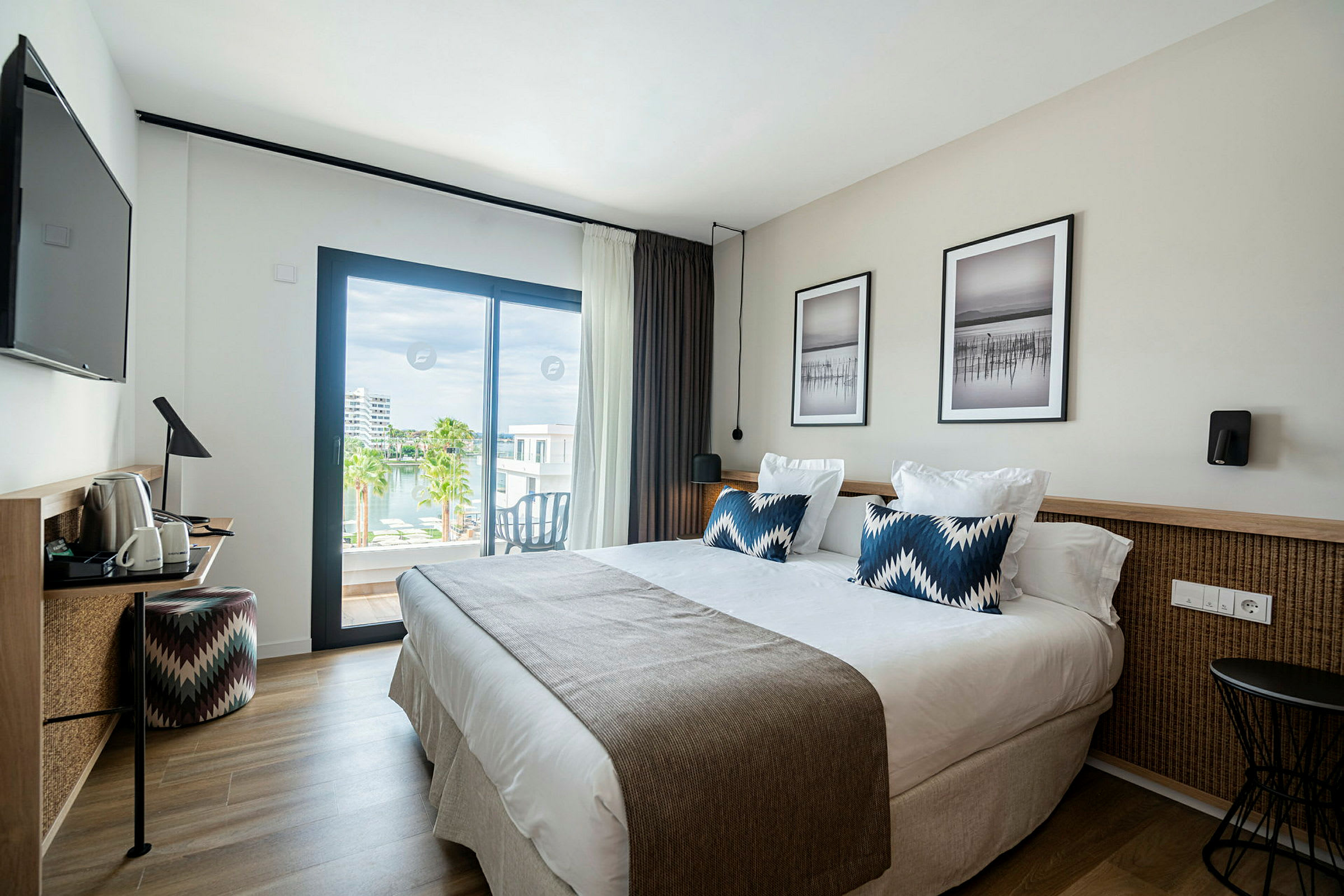 Et moderne hotellrom med en dobbeltseng, to puter og et teppe, samt veggkunst over sengen. Rommet har en skrivebordskrok med en stol og en TV på veggen. En stor glassdør fører ut til en balkong med utsikt over vannet og palmer. (AI-generert tekst)