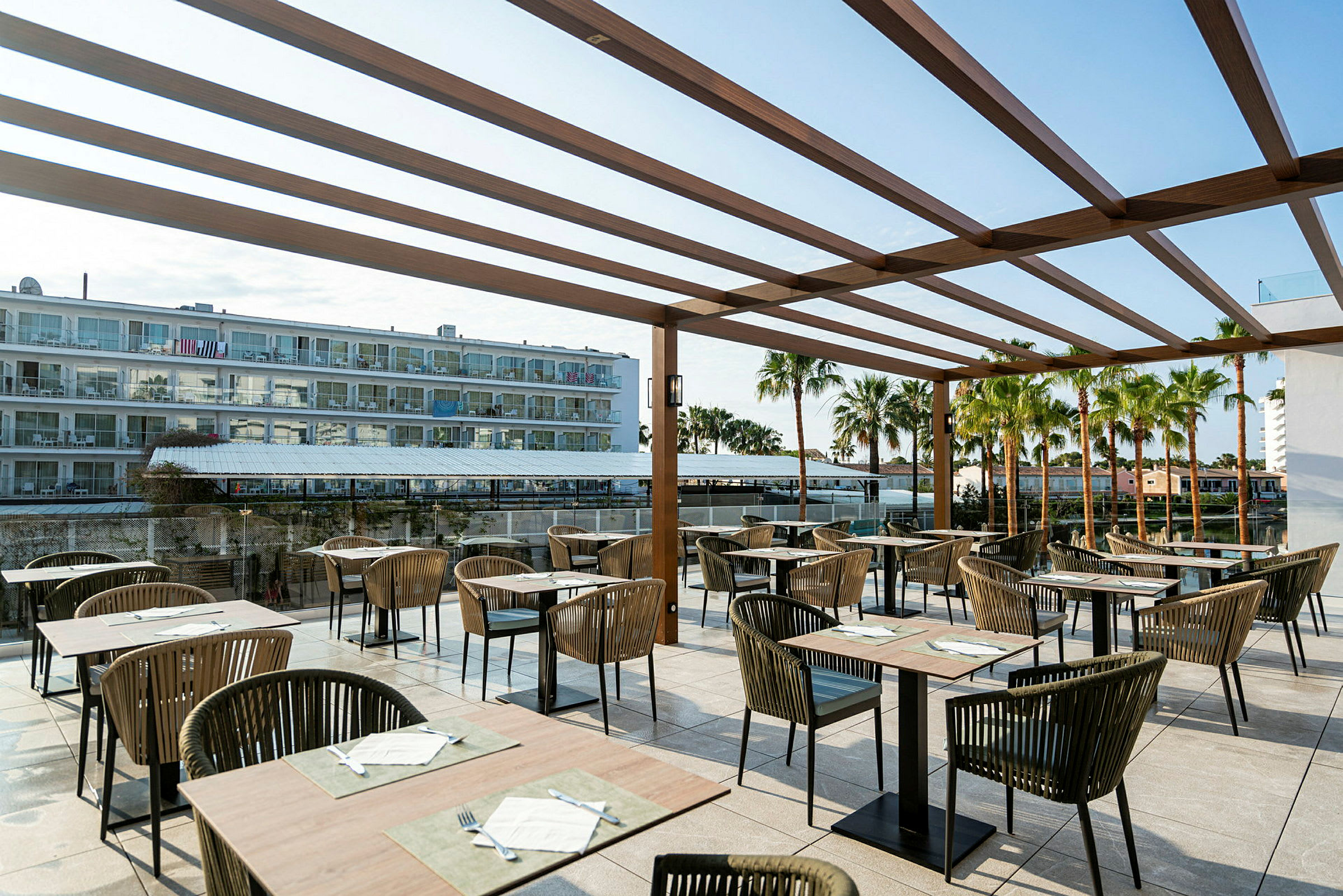 Bilde av en restaurantterrasse med bord og stoler under en trepergola. I bakgrunnen sees et hotell og palmer. (AI-generert tekst)