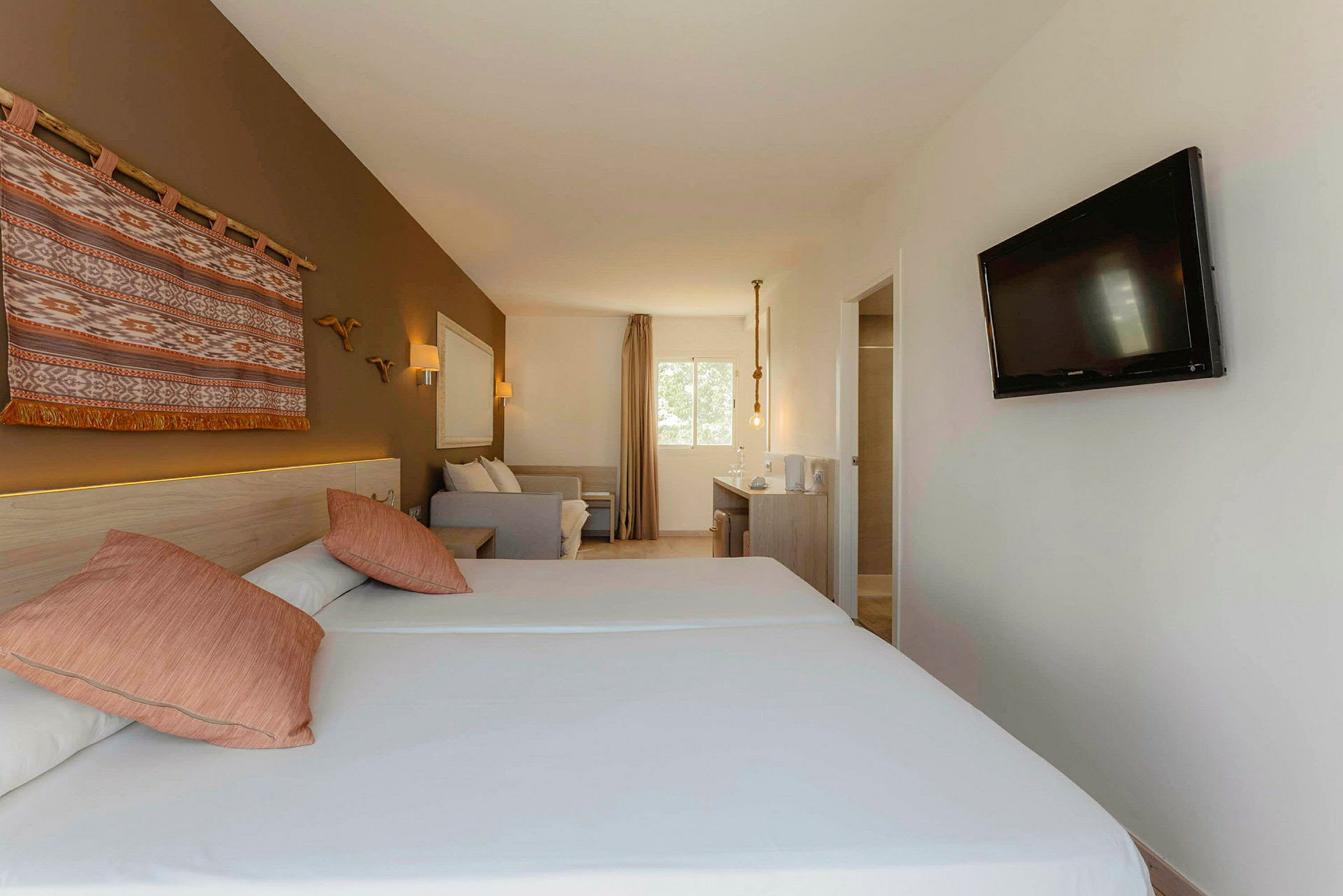 Et hotellrom med to enkeltsenger, en dekorativ veggteppe, en sofa, et skrivebord og en TV montert på veggen. (AI-generert tekst)