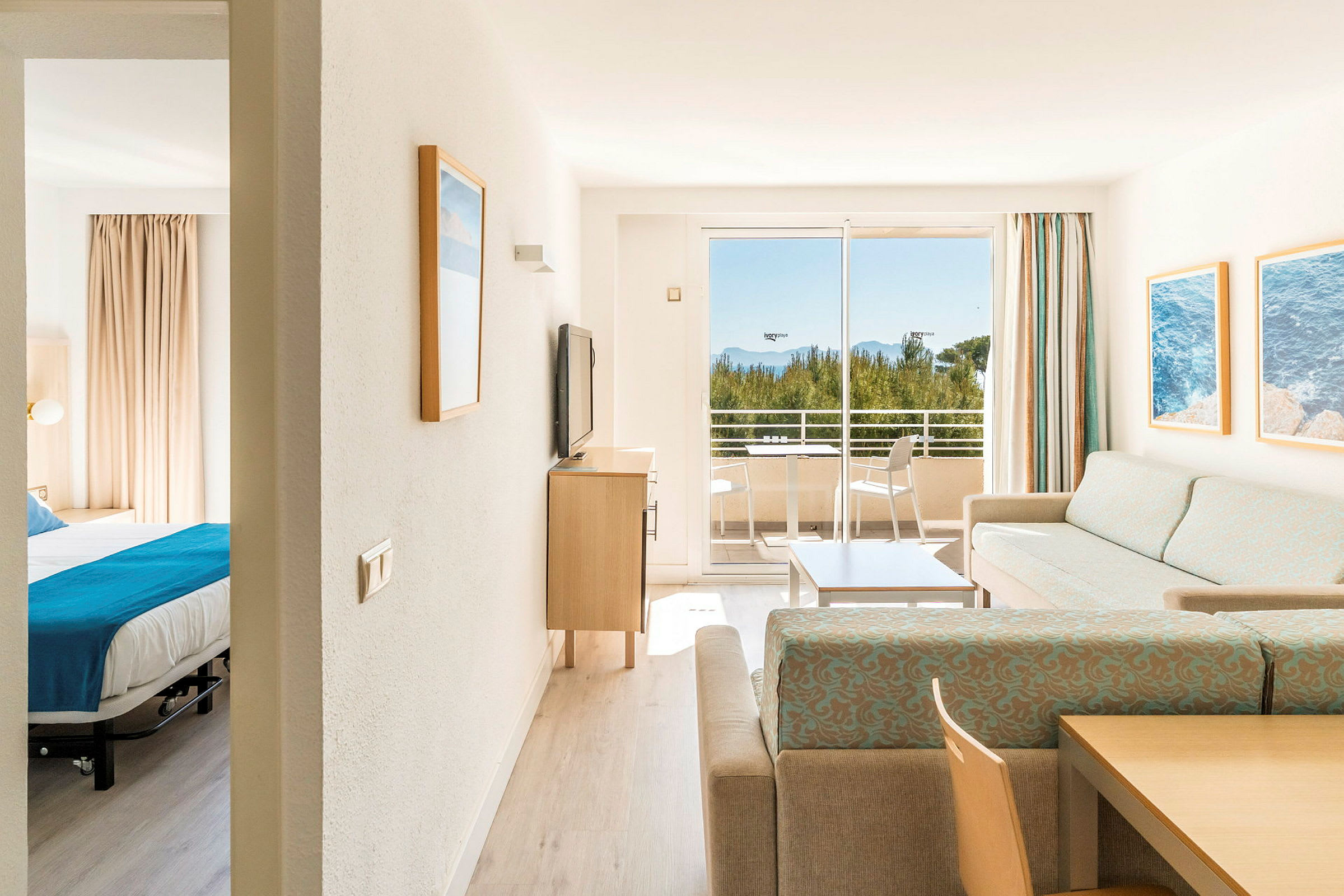 En lys hotellsuite med en stue, et soverom og en balkong som har utsikt over trær og hav. (AI-generert tekst)
