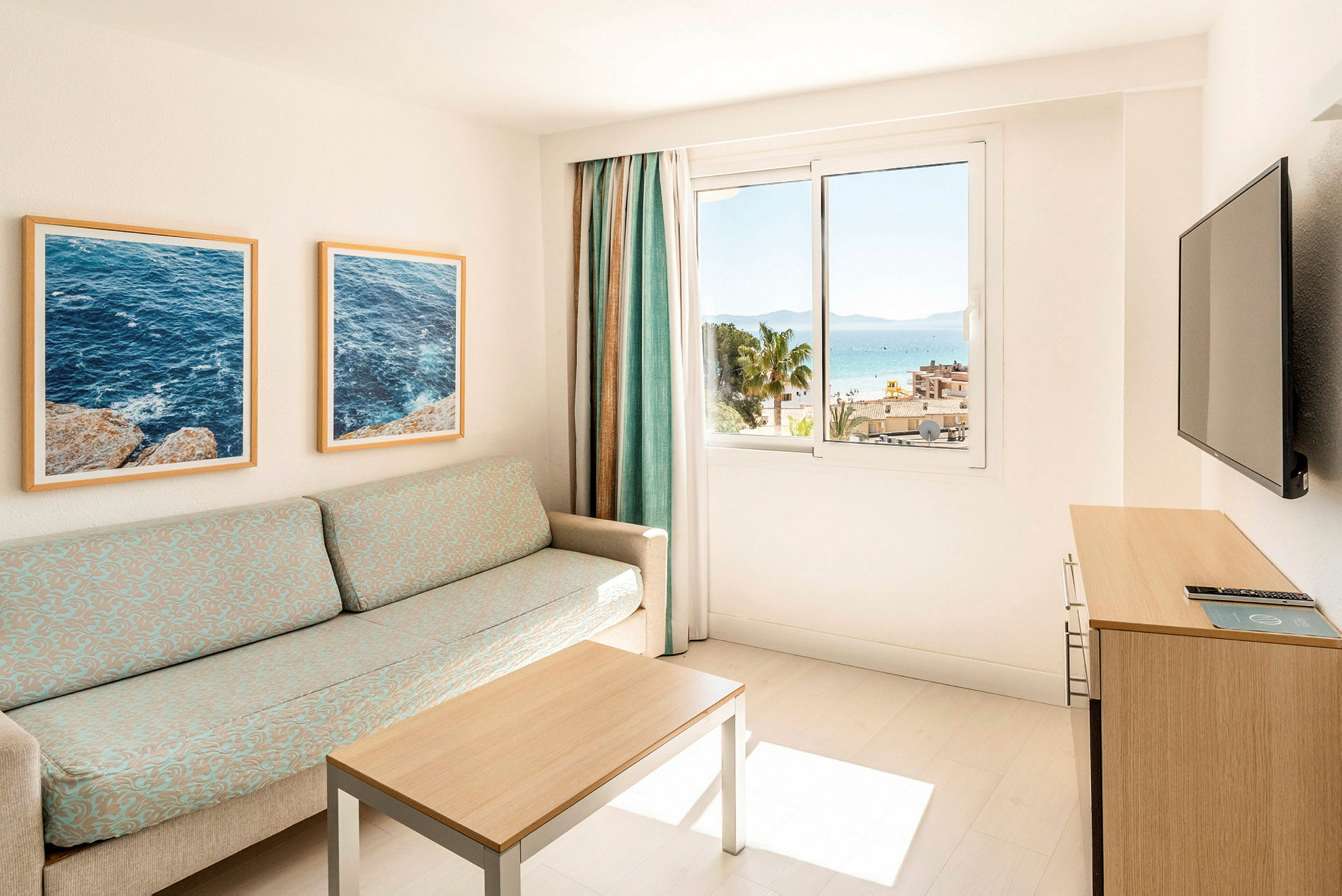 Et lyst hotellrom med en sofa, bord og en TV. Vinduet viser en utsikt over havet og palmer. (AI-generert tekst)