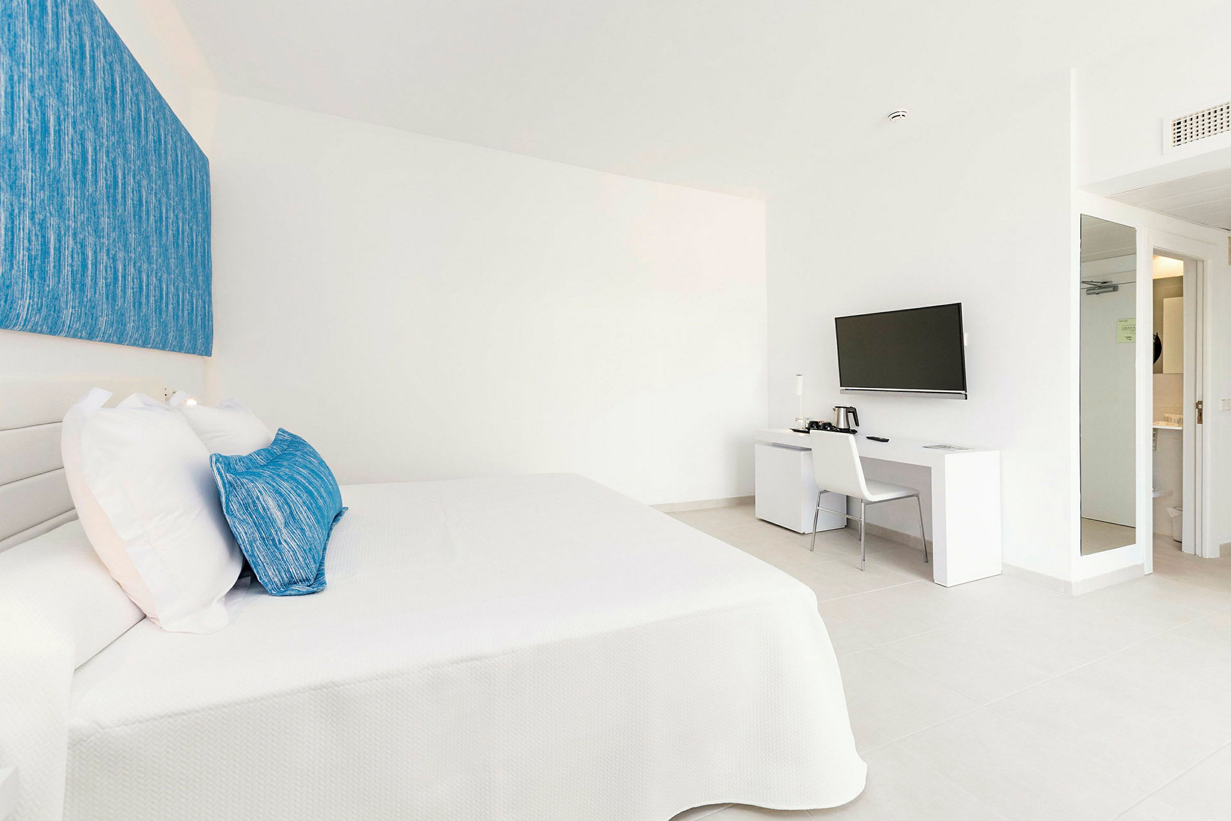 Et hotellrom med en hvit seng med blå puter, og et tilstøtende skrivebord med en veggmontert TV over. (AI-generert tekst)