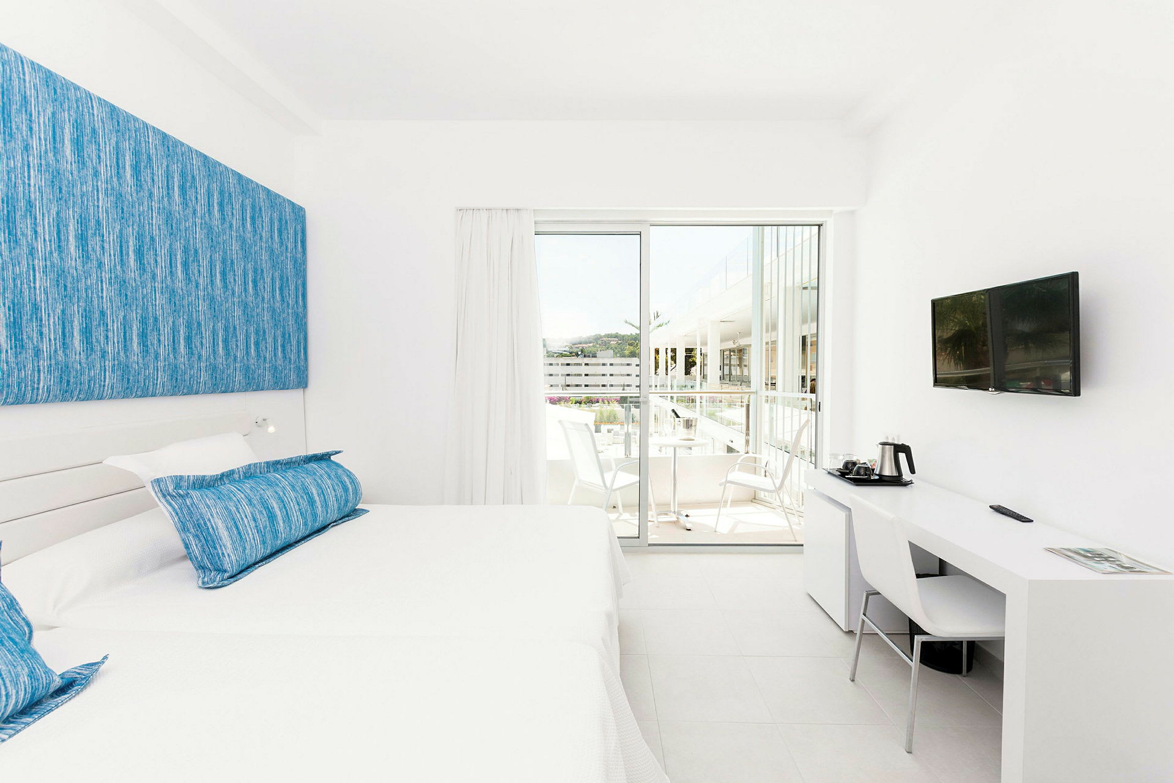 Et lyst hotellrom med hvit innredning, to enkeltsenger med blå puter og et skrivebord med stol og TV på veggen. En skyvedør leder ut til balkongen med sittemøbler. (AI-generert tekst)