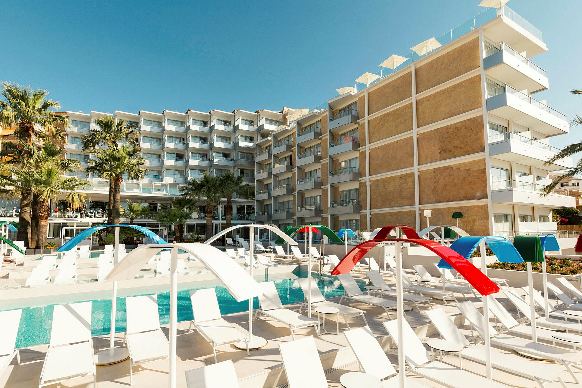 Et hotell med flere etasjer i moderne design er synlig bak et bassengområde med solsenger og fargerike solskjermer. (AI-generert tekst)