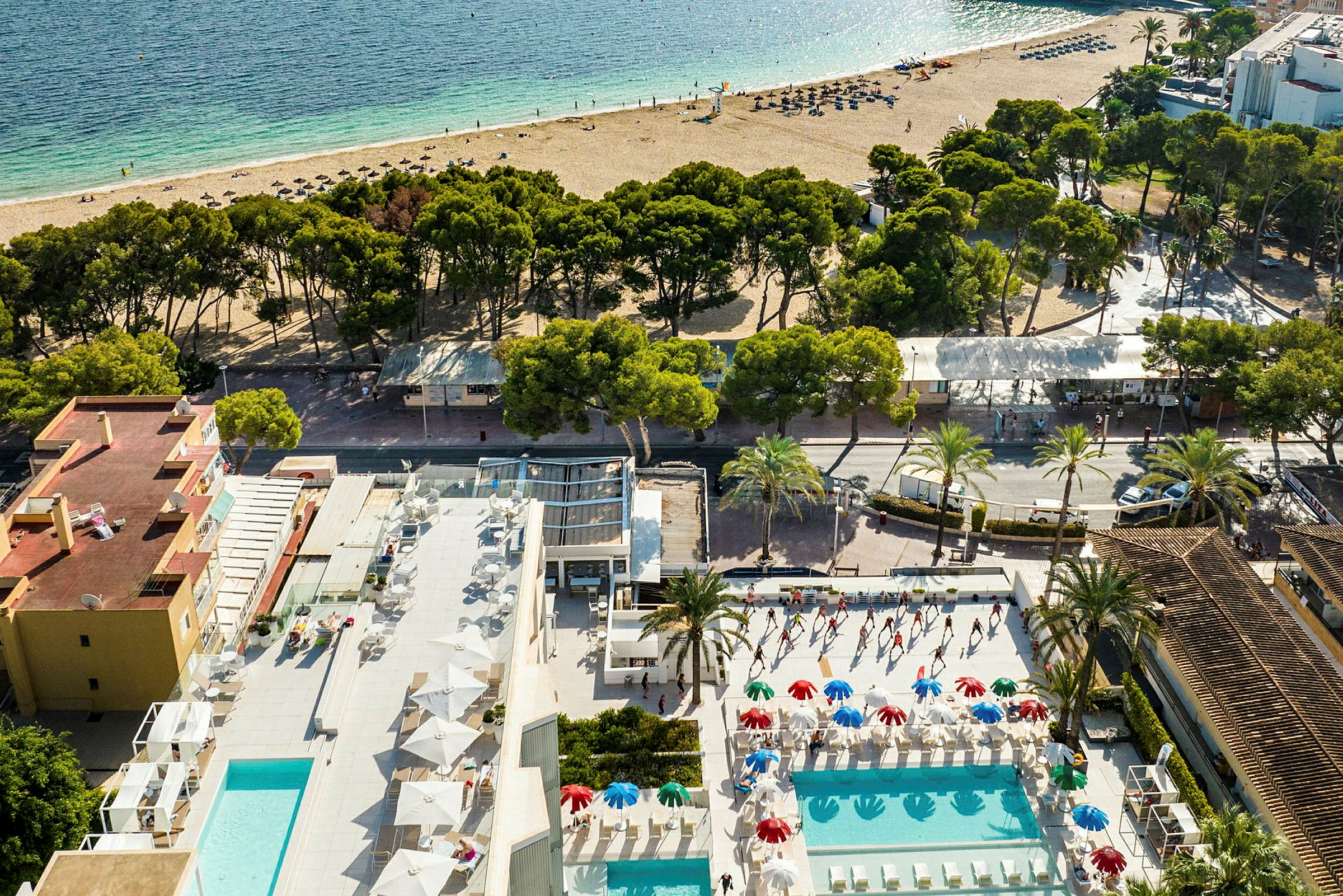 Utsikt ovenfra av et hotellområde med utendørsbassenger, parasoller og solsenger. En sandstrand og havet er synlig i bakgrunnen. (AI-generert tekst)