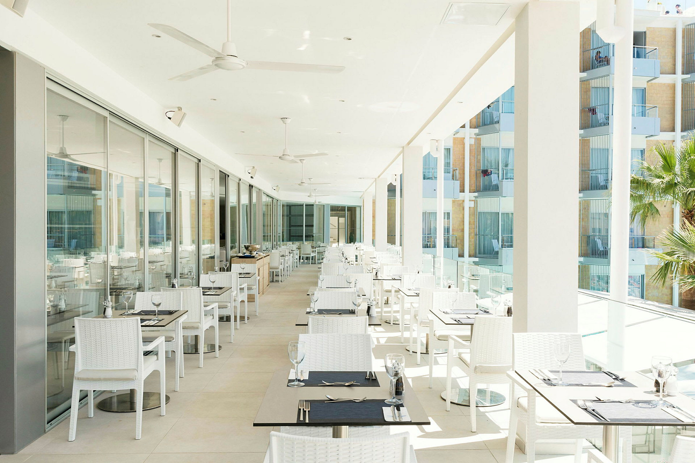 En lyst restaurantterrasse med mange hvite bord og stoler, glassdører langs den ene siden og utsikt over balkonger utendørs. (AI-generert tekst)