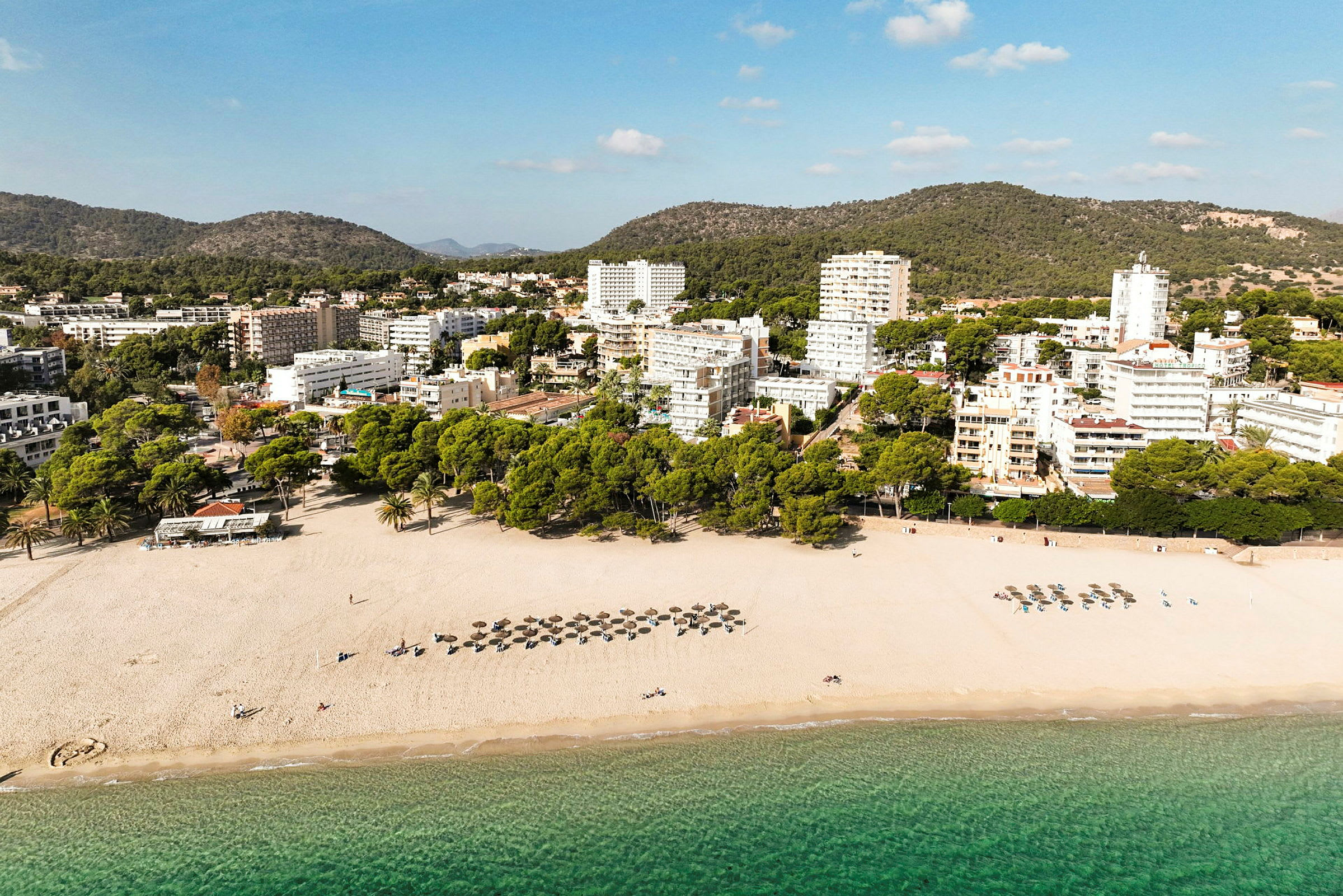 Strand med parasoller og solsenger ved klart vann, bak en rad trær og bygninger. (AI-generert tekst)