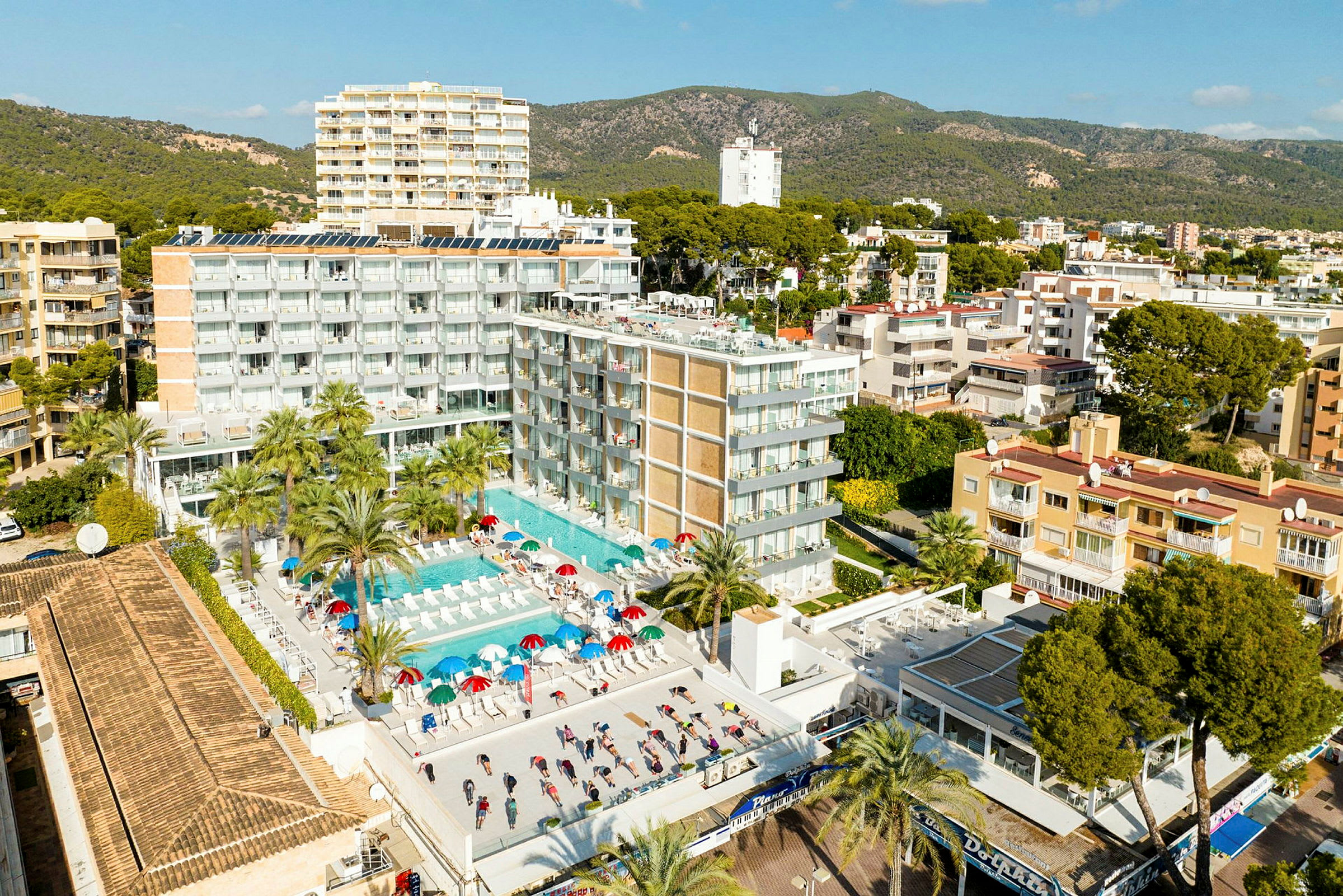 Flyfoto av en hotellbygning med bassengområde og solsenger. Omgivende landskap med grøntområder og flere bygninger i bakgrunnen. (AI-generert tekst)
