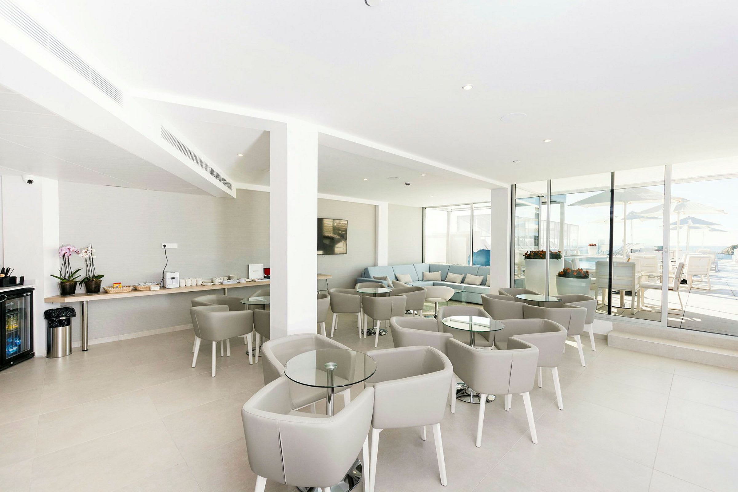 En moderne lounge med grå møbler og glassbord. En serveringsbenk med drikke og snacks er synlig til venstre, og store glassvinduer åpner opp til en utendørsterrasse. (AI-generert tekst)