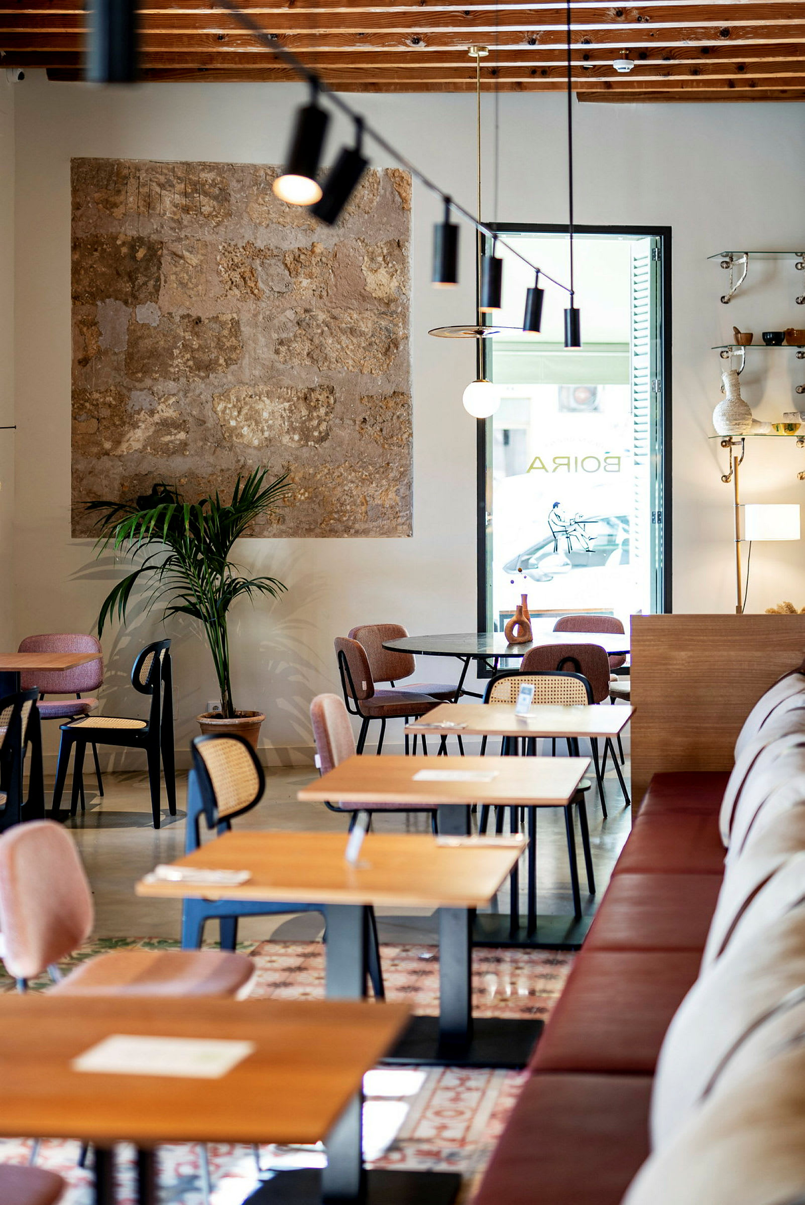 En moderne kafé med trebord og stoler samt en plante ved en vegg med stein. Lyset fra vinduet lyser opp rommet. (AI-generert tekst)