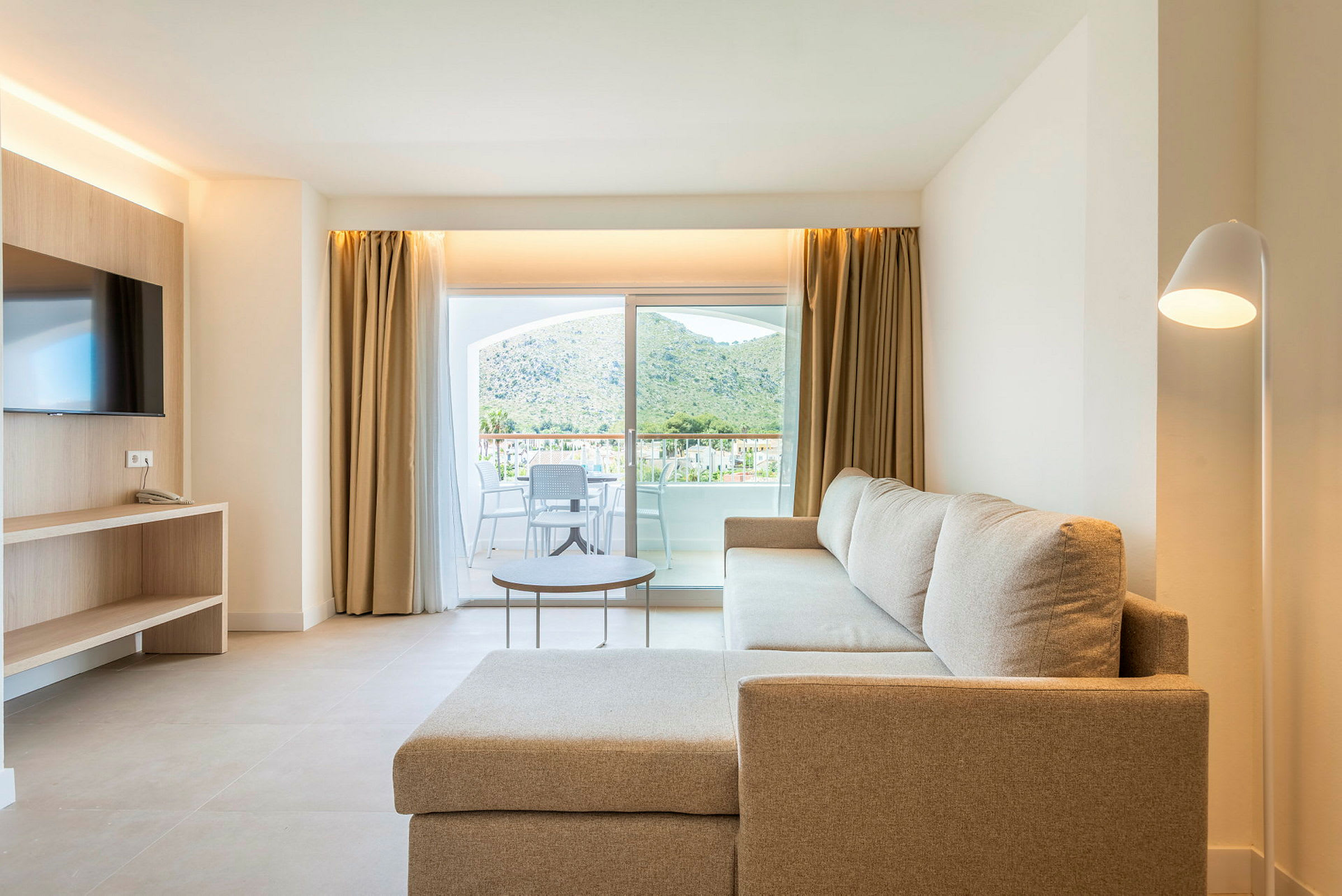 Stueinteriør med beige sofa, TV og glassdør med utsikt mot en terrasse og fjell. (AI-generert tekst)