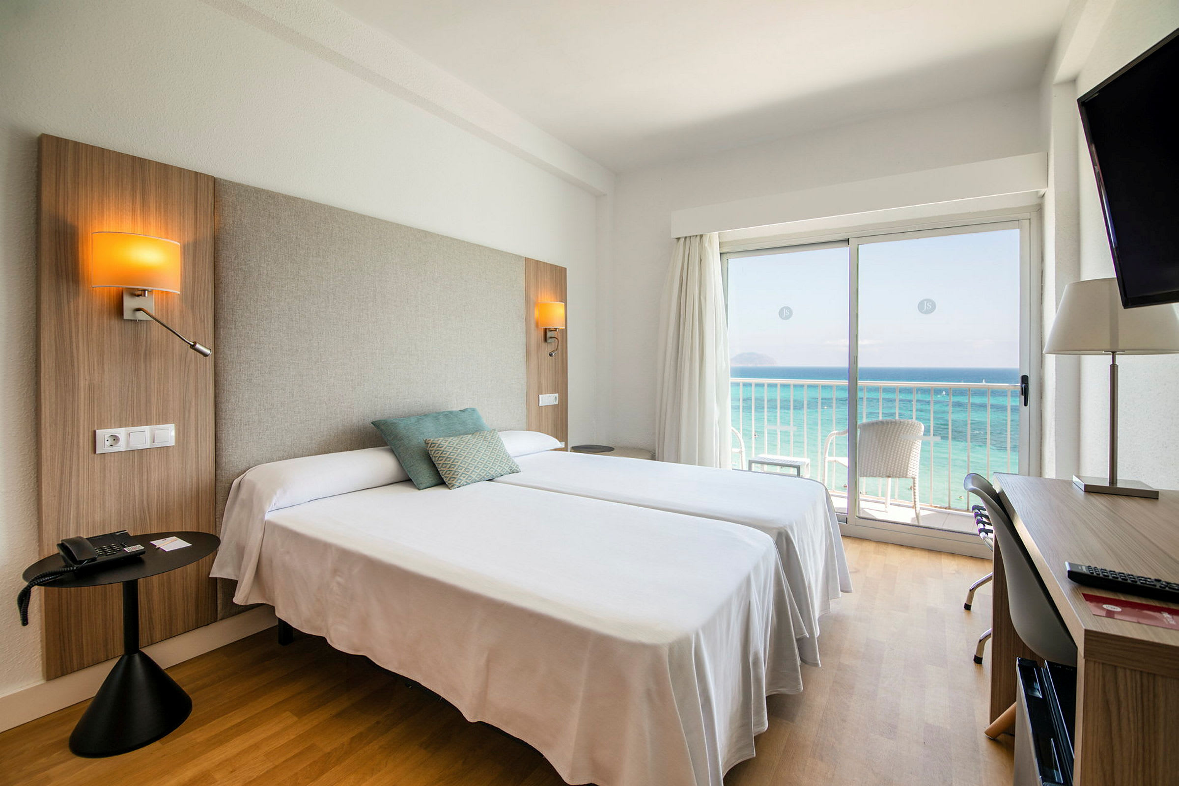 Hotellrom med to senger, havutsikt gjennom en glassdør og skrivebord. (AI-generert tekst)