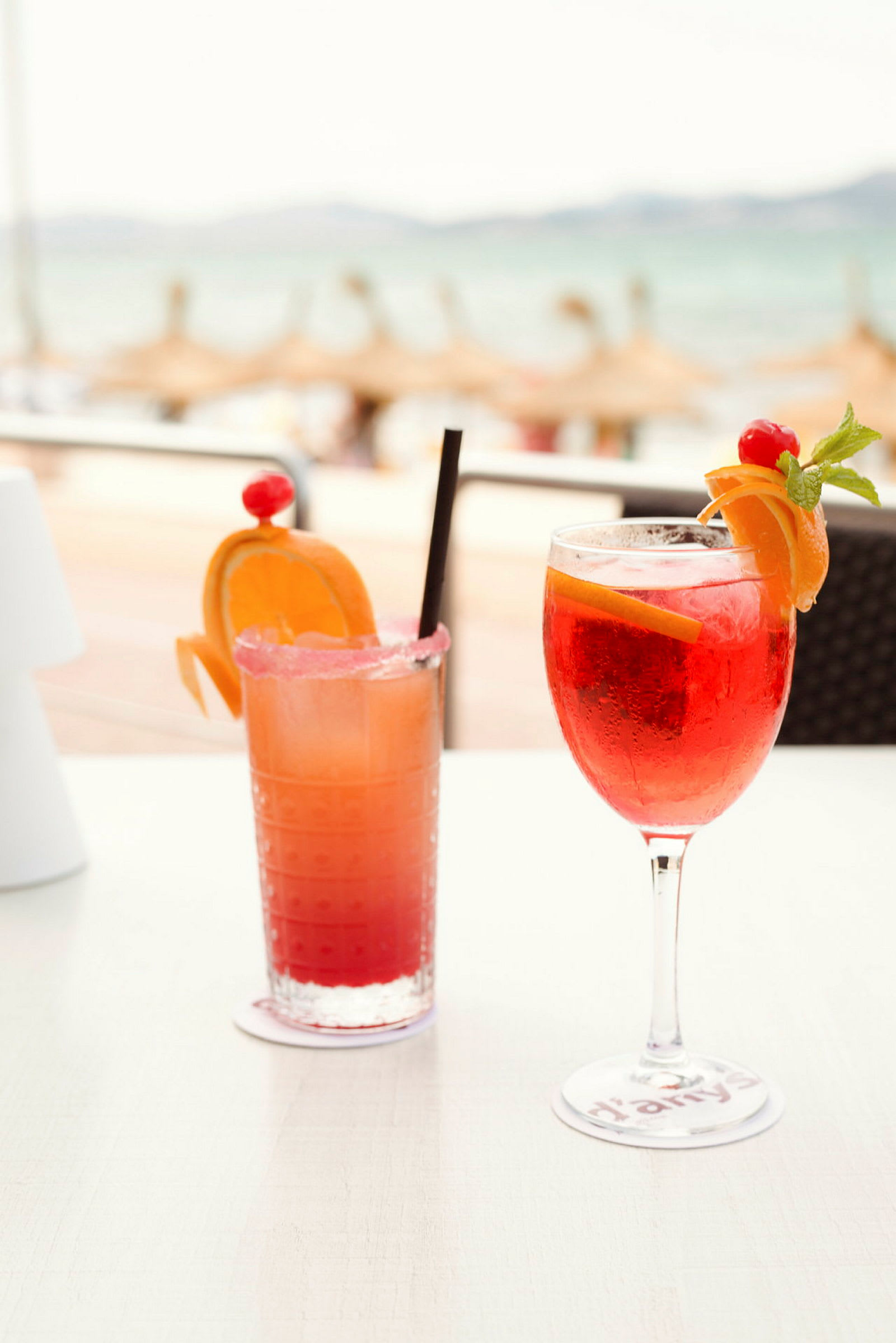 To fargerike drinker dekorert med frukt på et bord, med en uskarp strand i bakgrunnen. (AI-generert tekst)