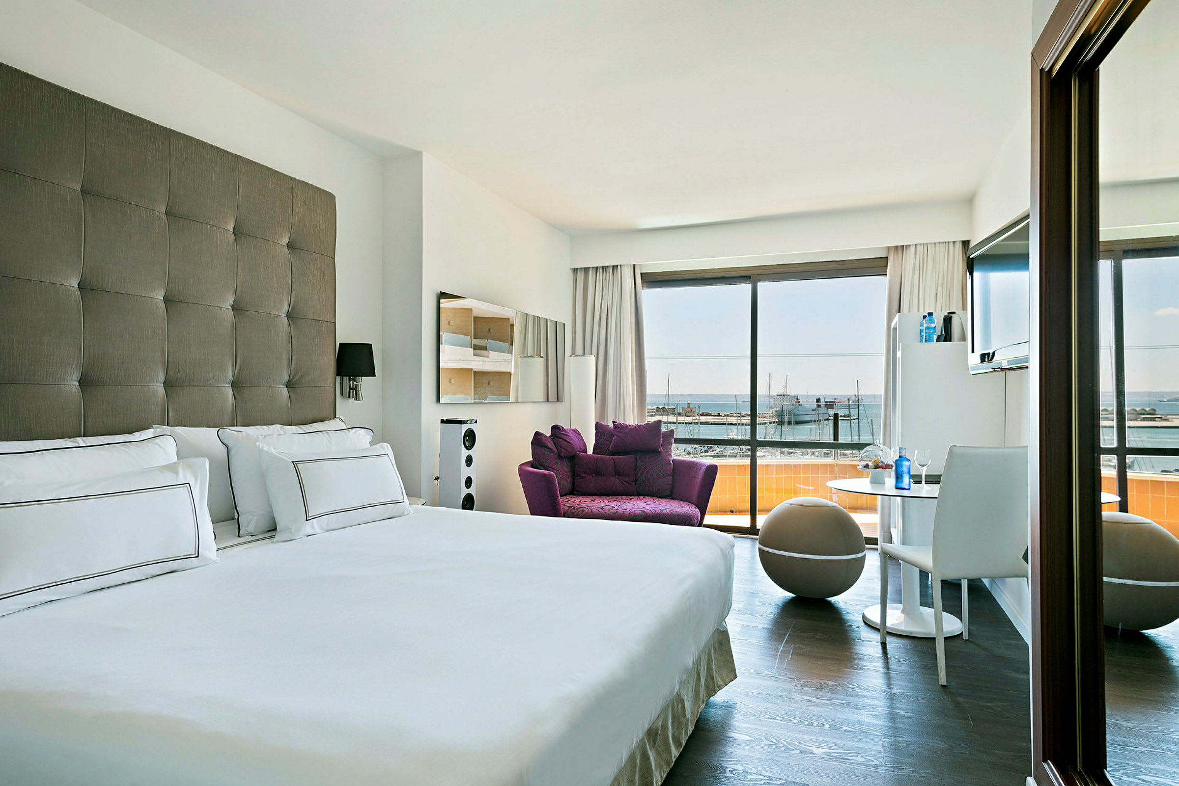 Et moderne innredet hotellrom med en dobbeltseng, en lilla lenestol og utsikt over en marina gjennom store glassvinduer. (AI-generert tekst)