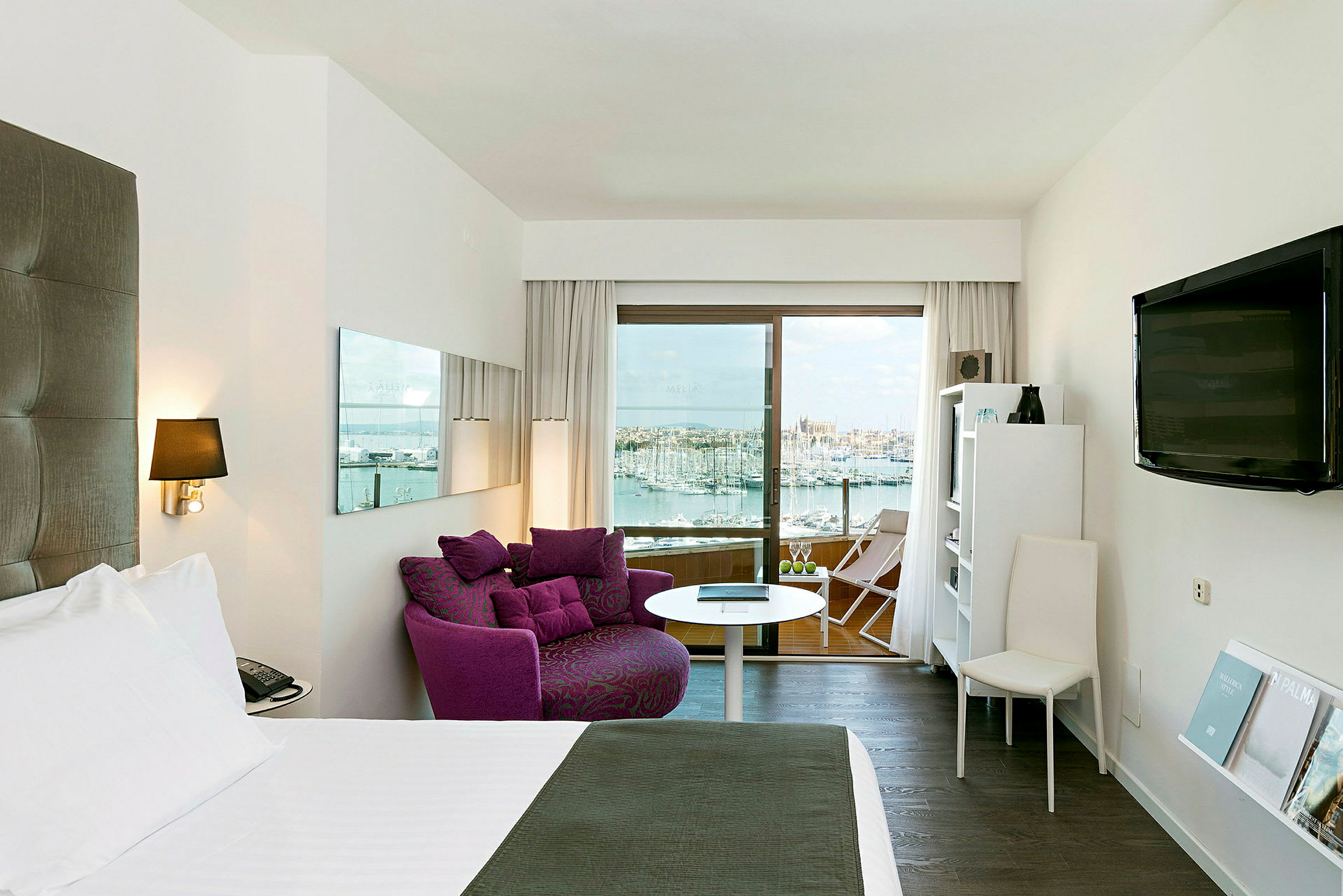 Hotellrom med en bred seng, lilla lenestol, en TV på veggen og en balkong med utsikt over havnen og byen. (AI-generert tekst)