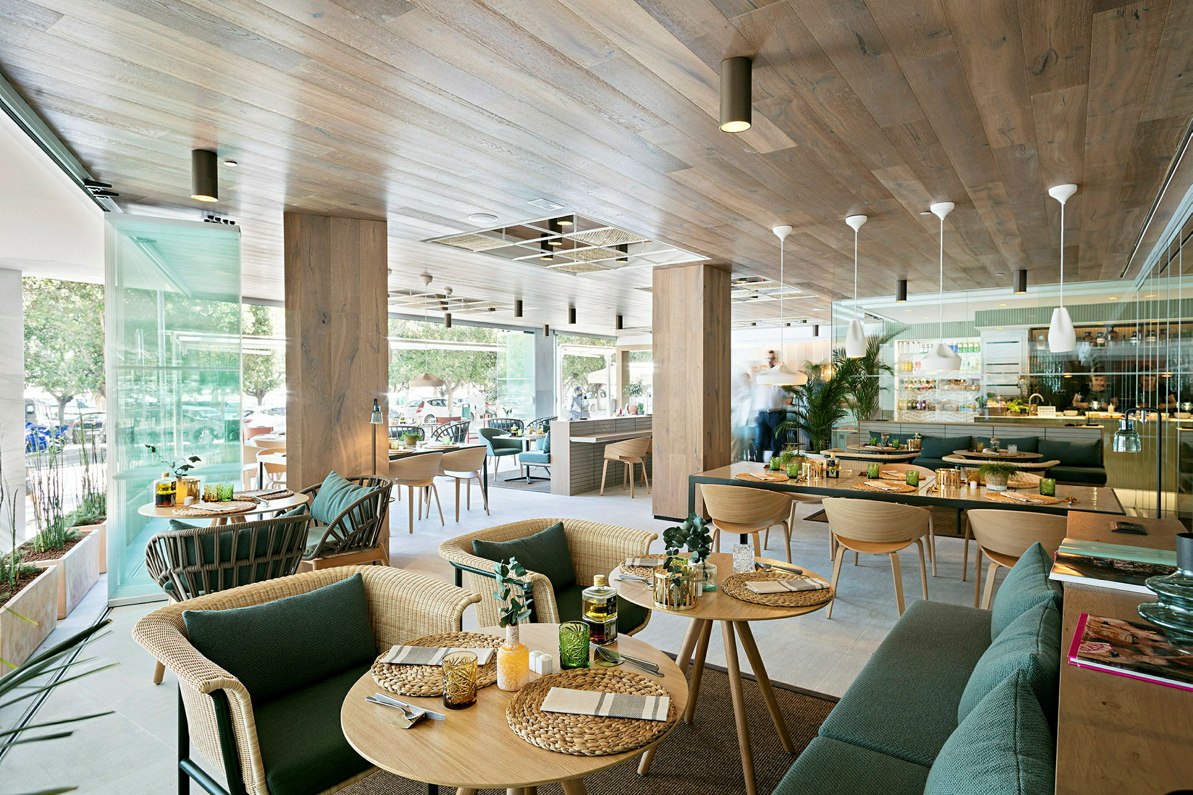 En moderne restaurant med treinteriør og store glassvinduer. Det er bord med servert mat og interiør med grønne planter. (AI-generert tekst)