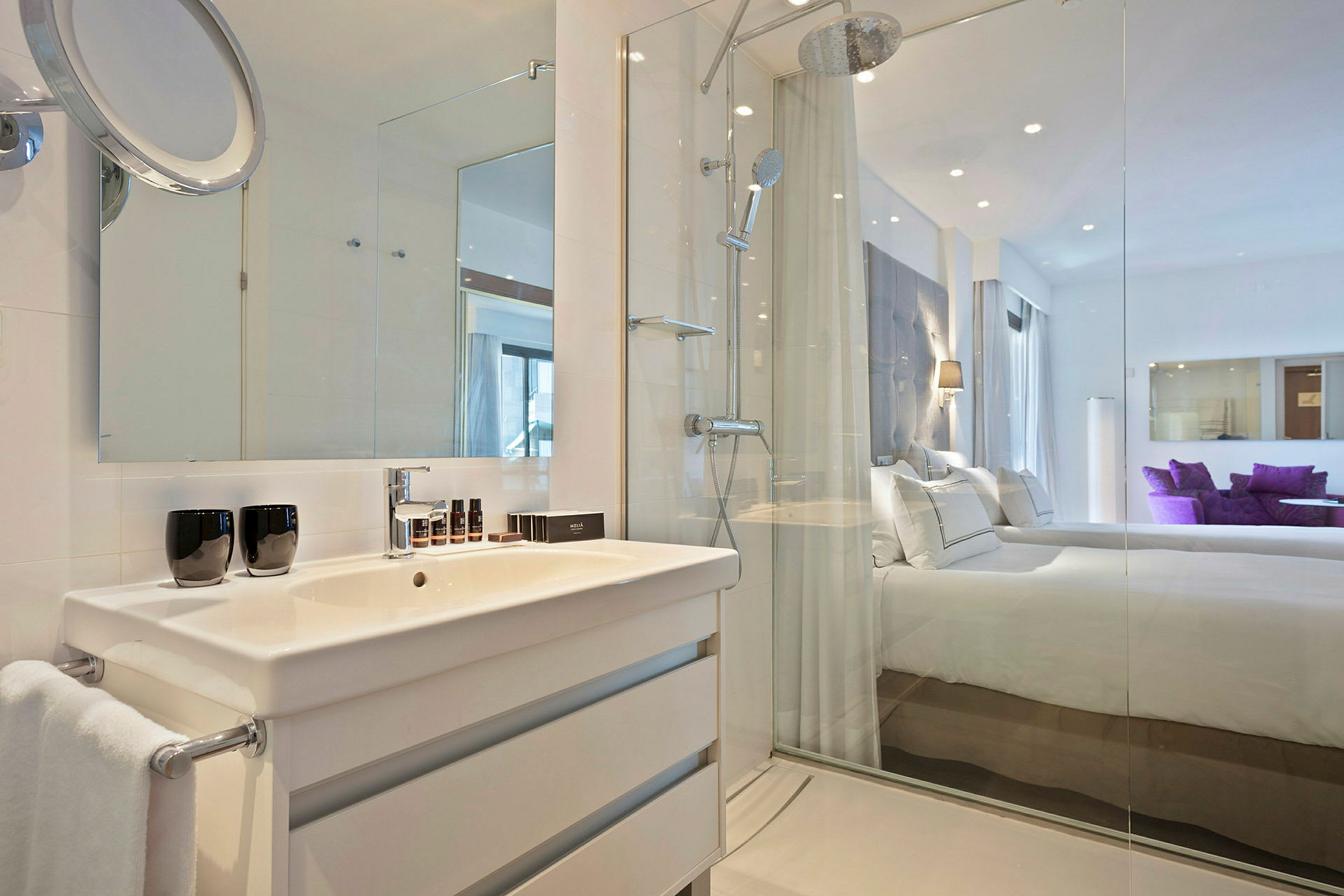 Et moderne bad med hvite møbler, servant og dusj. En glassvegg skiller badet fra et soverom som ses i bakgrunnen. (AI-generert tekst)