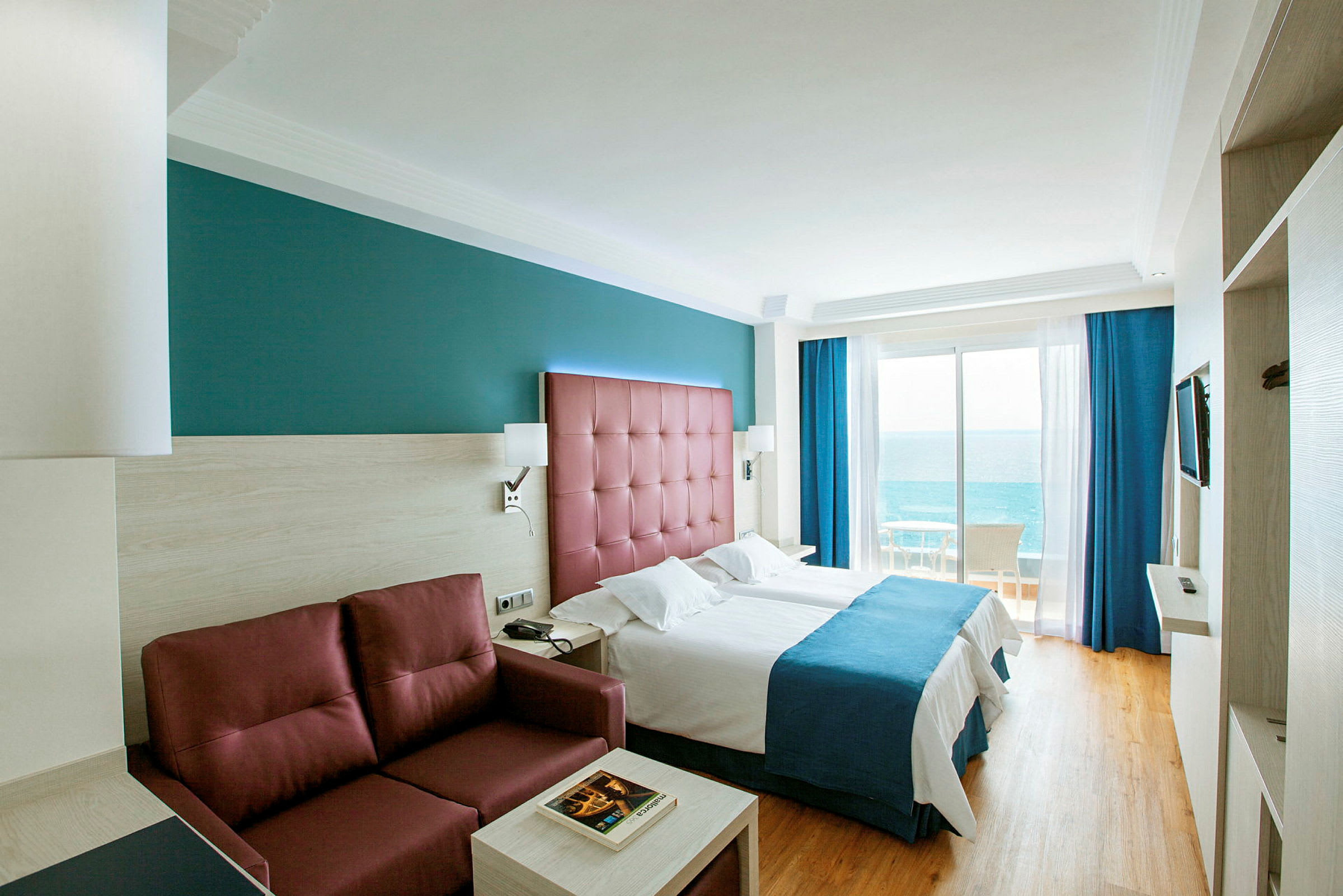 Hotellrom med to enkeltsenger, en rød sofa og havutsikt fra balkongen. (AI-generert tekst)