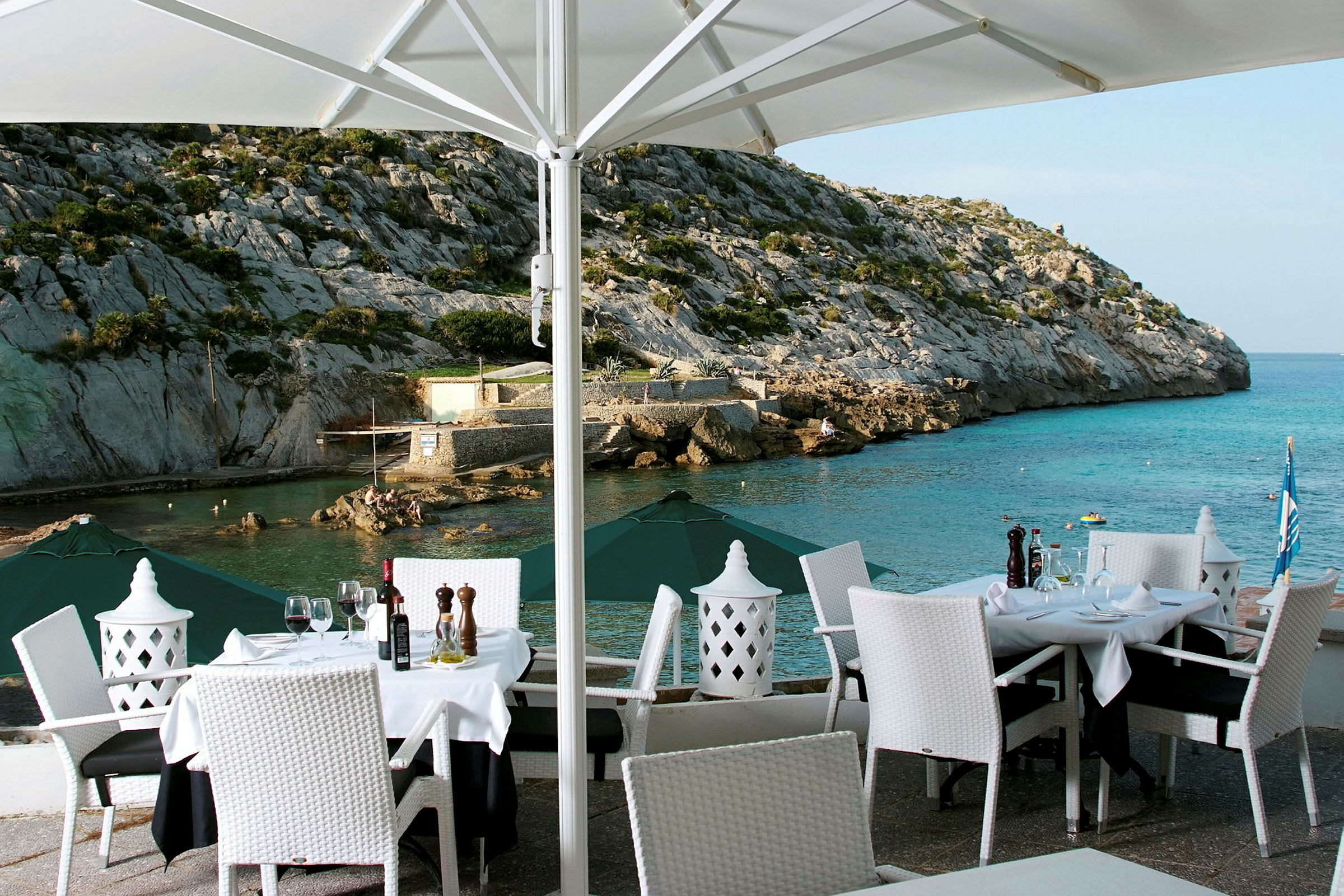 En utendørs restaurant med dekkede bord og hvite stoler med utsikt over et steinete landskap og havet. (AI-generert tekst)