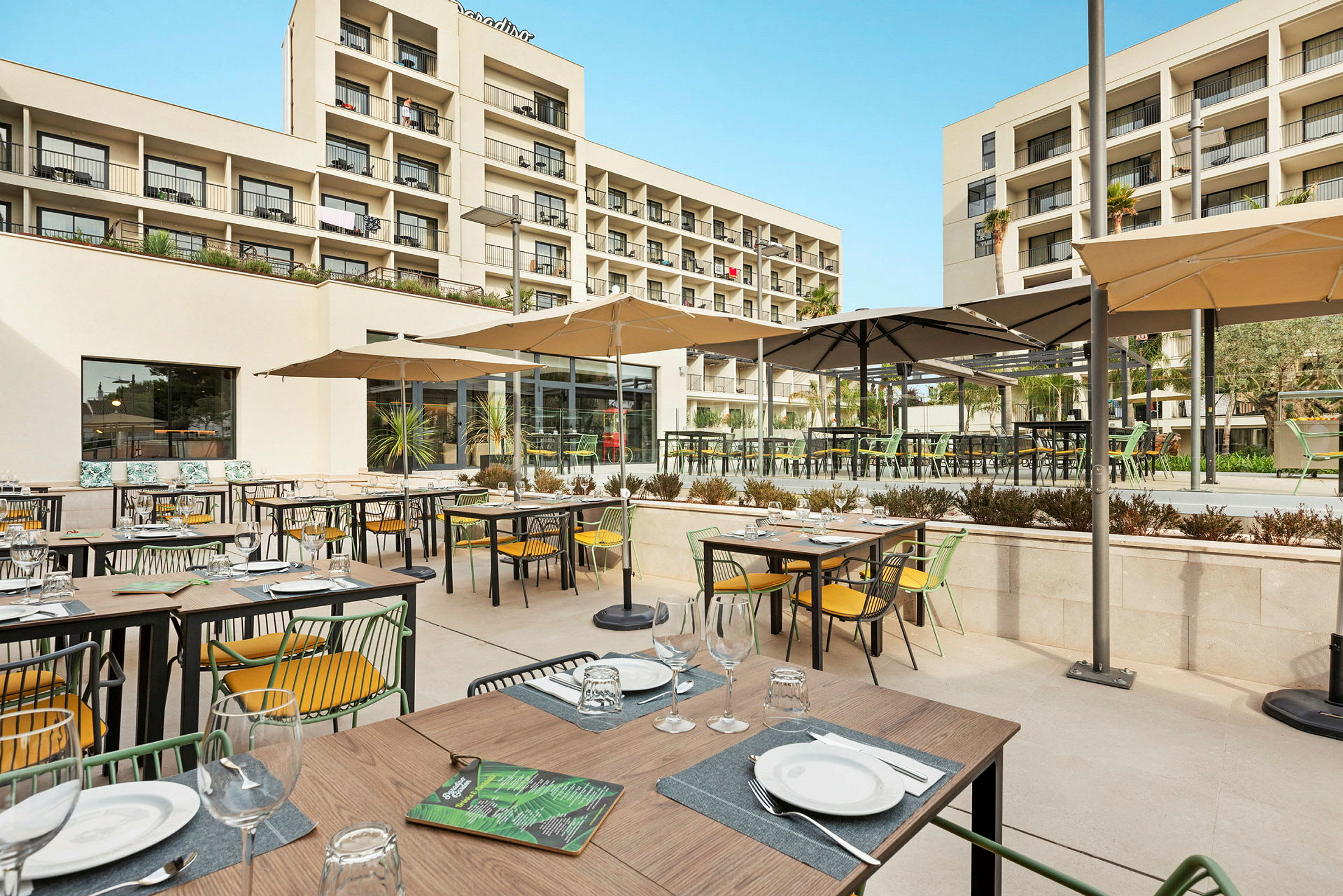 Bilde av en utendørsrestaurant med oppstilte bord og stoler under parasoller, samt hotellbygningen i bakgrunnen. (AI-generert tekst)