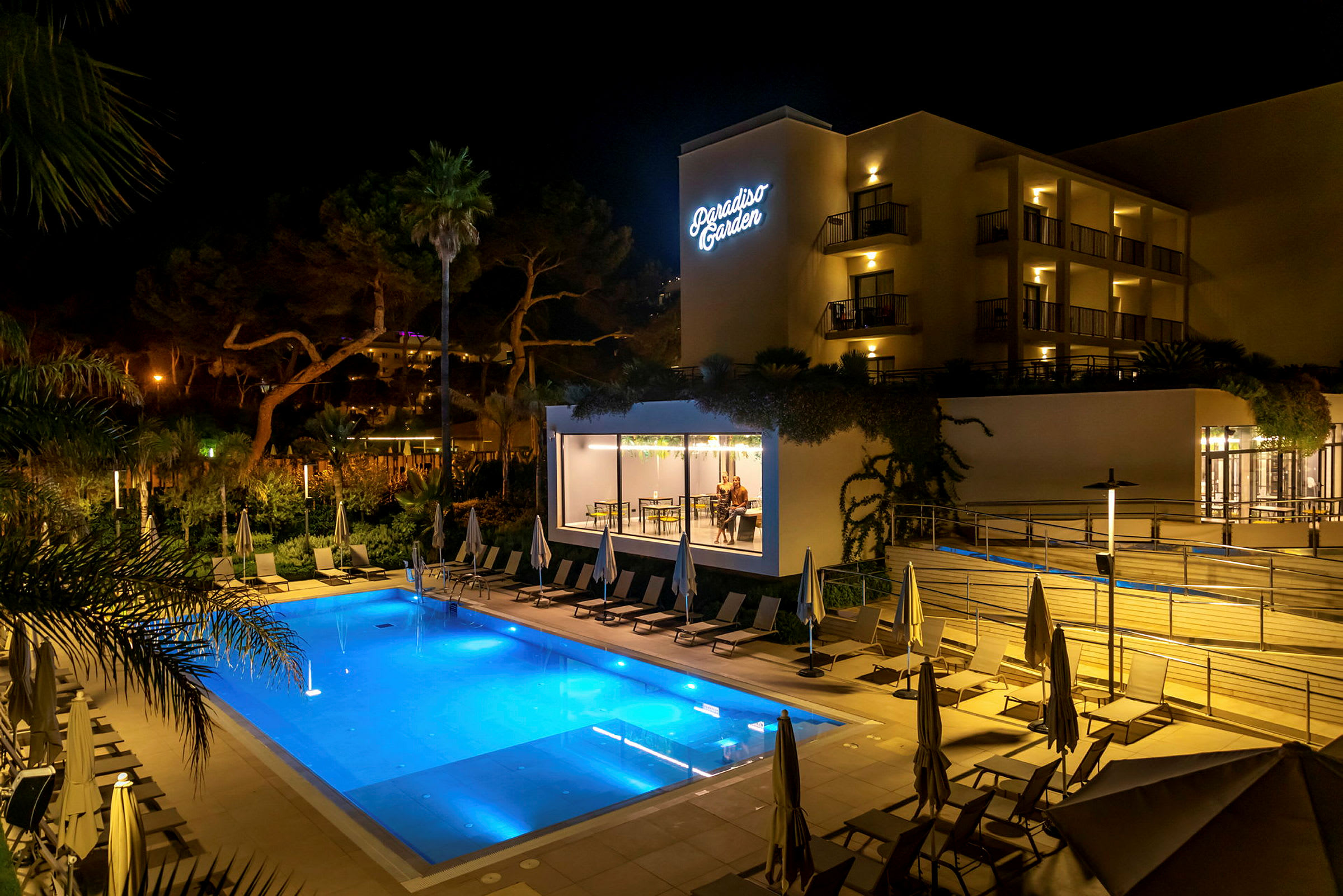 Hotell Paradiso Garden sett om kvelden med et opplyst basseng og solsenger i forgrunnen. (AI-generert tekst)