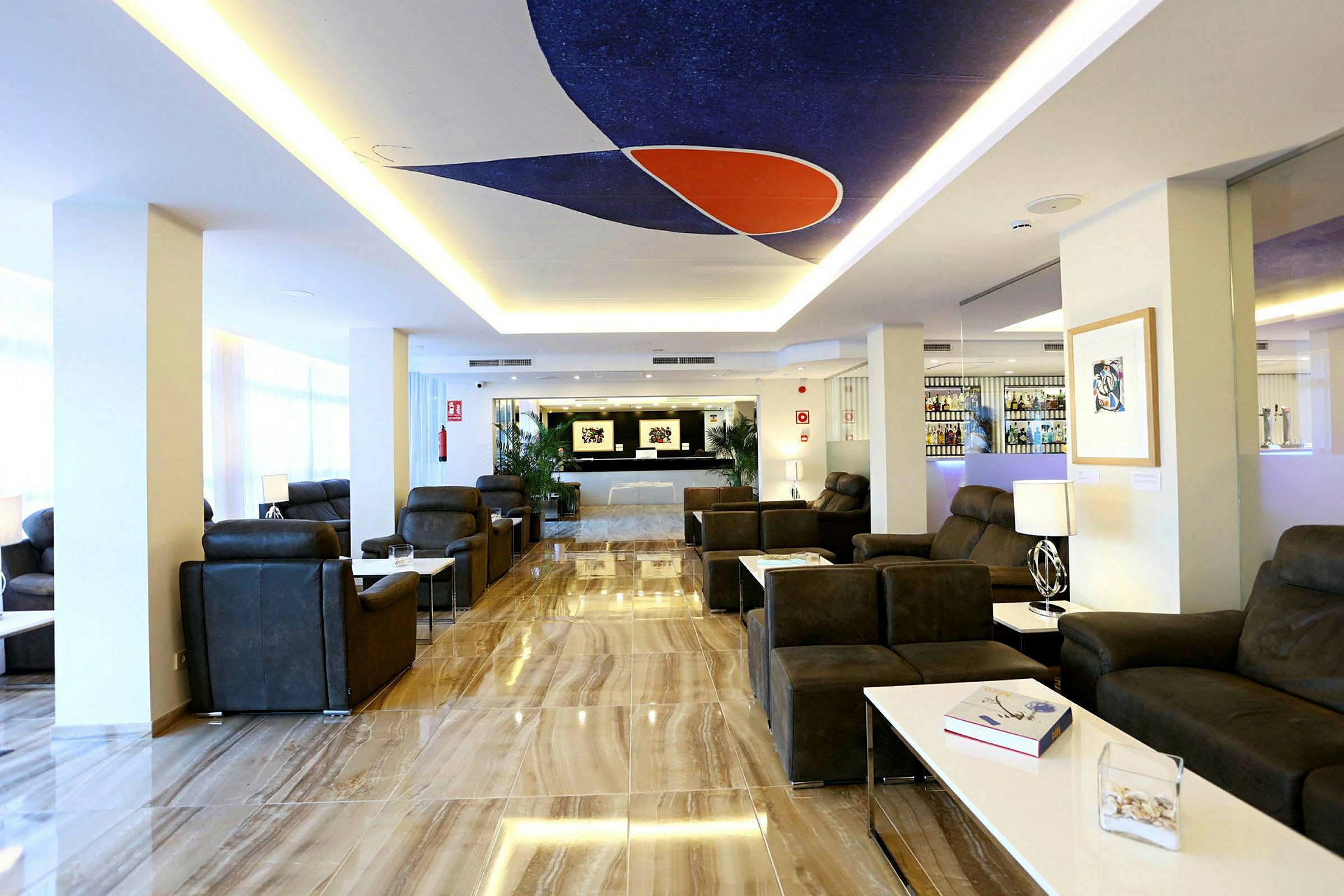 En moderne innredet hotellobby med mørke sofaer, hvite bord og et fargerikt dekorativt panel i taket. (AI-generert tekst)