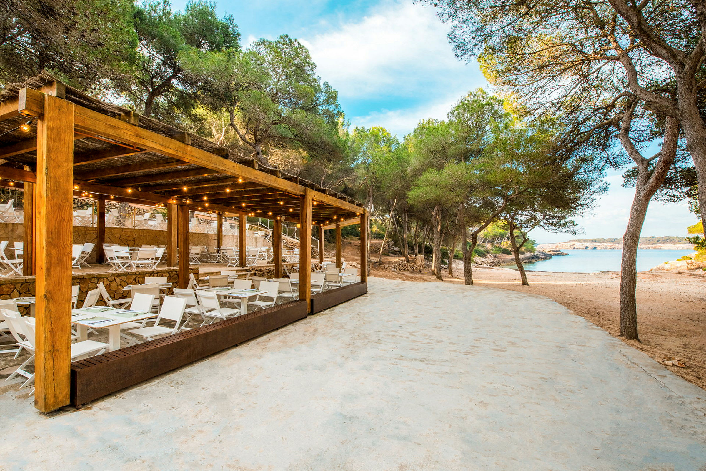 En strandrestaurant med hvite møbler og en trepergola nær sandstrand og trær. (AI-generert tekst)