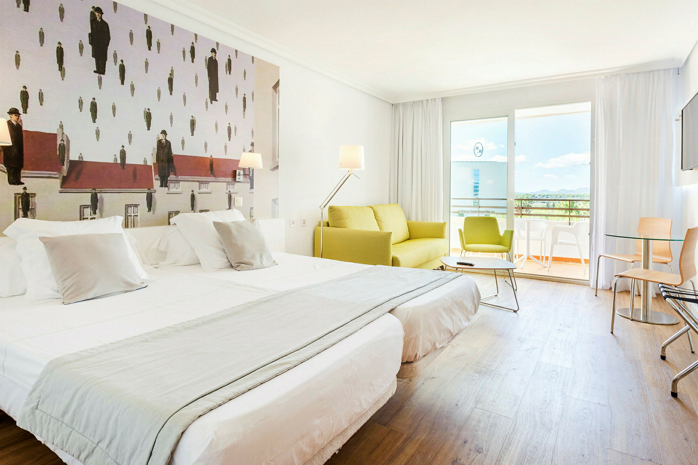 Et hotellrom med to enkelt oppredde senger, en grønn sofa og en balkong i bakgrunnen. Veggdekorasjonen har et kunstverk med uvanlig design. (AI-generert tekst)